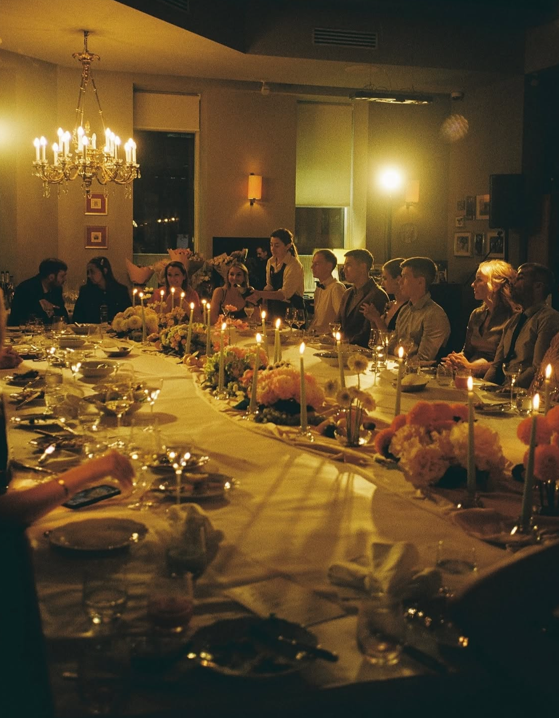 Groupe de personnes assises autour d'une longue table décorée pour un dîner, avec des bougies et des fleurs, dans une salle élégante avec un lustre suspendu.