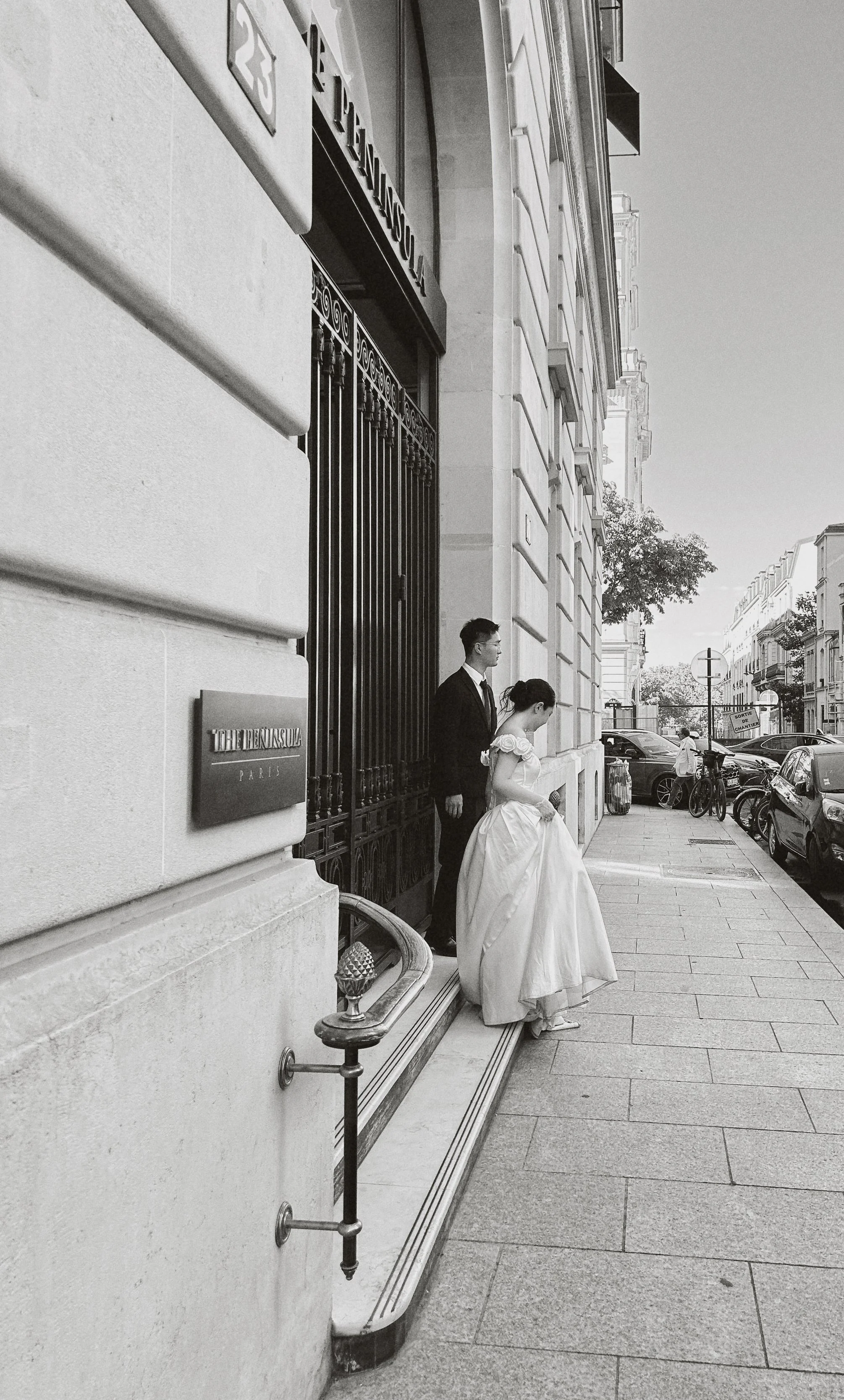 Deux personnes habillées en mariée et en costume debout à côté d'une porte dans une rue parisienne. La femme porte une robe de mariée blanche volumineuse et le homme un costume sombre. Le bâtiment a une façade en pierre avec une grille noire devant la porte.