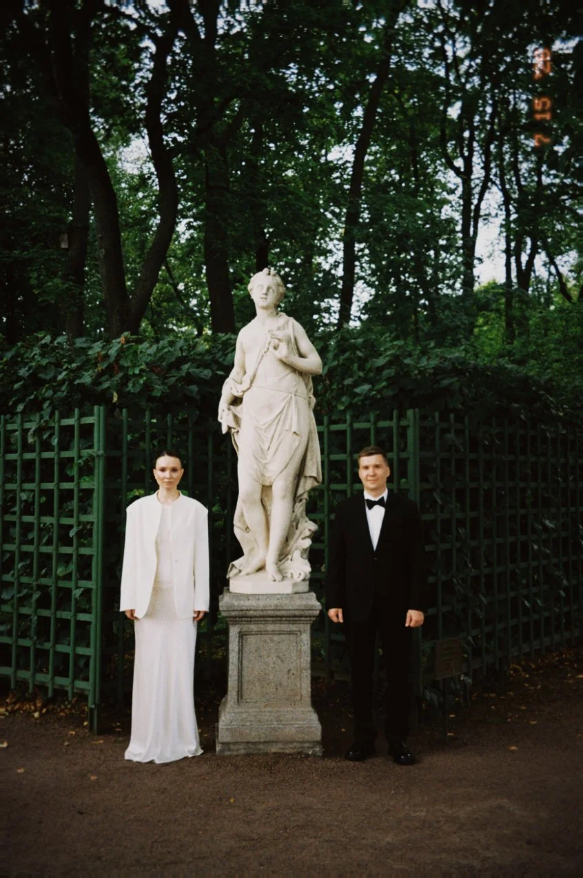 Un homme et une femme en tenue formelle se tiennent à côté d'une statue en pierre dans un parc verdoyant. La femme porte une robe longue blanche et le homme un costume noir avec une cravate noire. Le fond est constitué d'un arboré dense avec un panneau de clôture en métal vert.