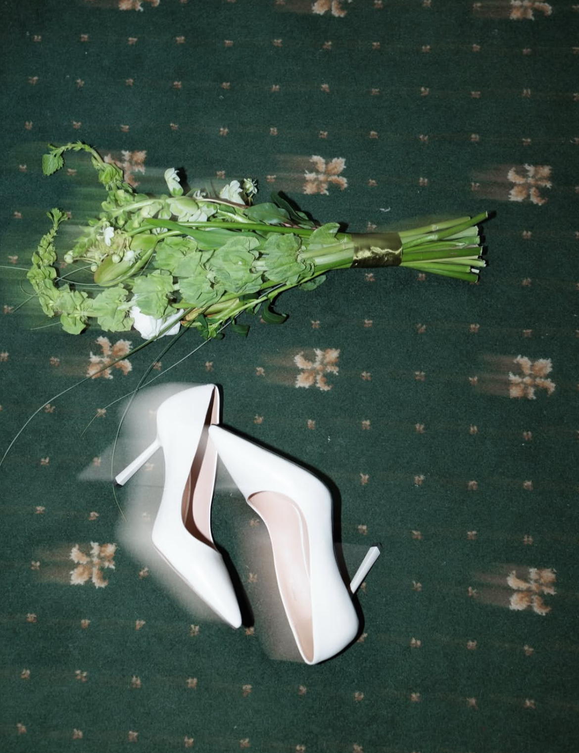 Une paire de chaussures à talons hauts blanches sur un tapis vert avec un motif floral, devant une bouquet de fleurs vertes et blanches.