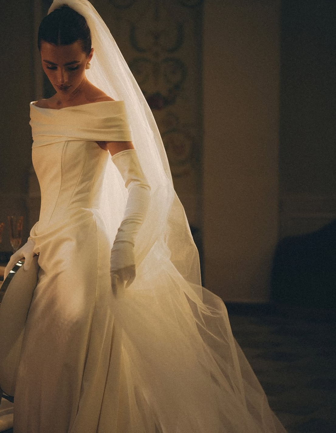 Une femme en robe de mariée blanche et longue, portant des gants longs, dans un intérieur sombre.