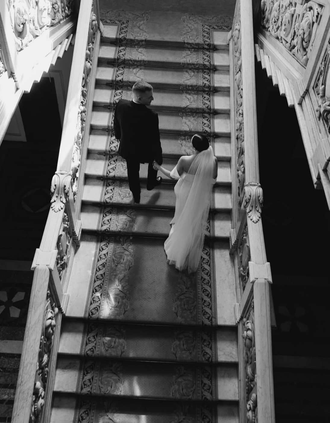 Un couple en mariage, avec la femme en robe blanche et voile, montant un escalier en marbre décoré, main dans la main dans un endroit élégant.