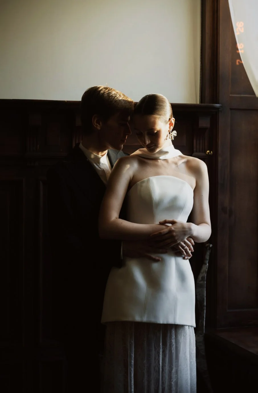 Un couple en tenue de mariage, la femme porte une robe blanche et l'homme un costume noir, partage un moment intime dans une pièce sombre avec une lumière naturelle venant d'une fenêtre.