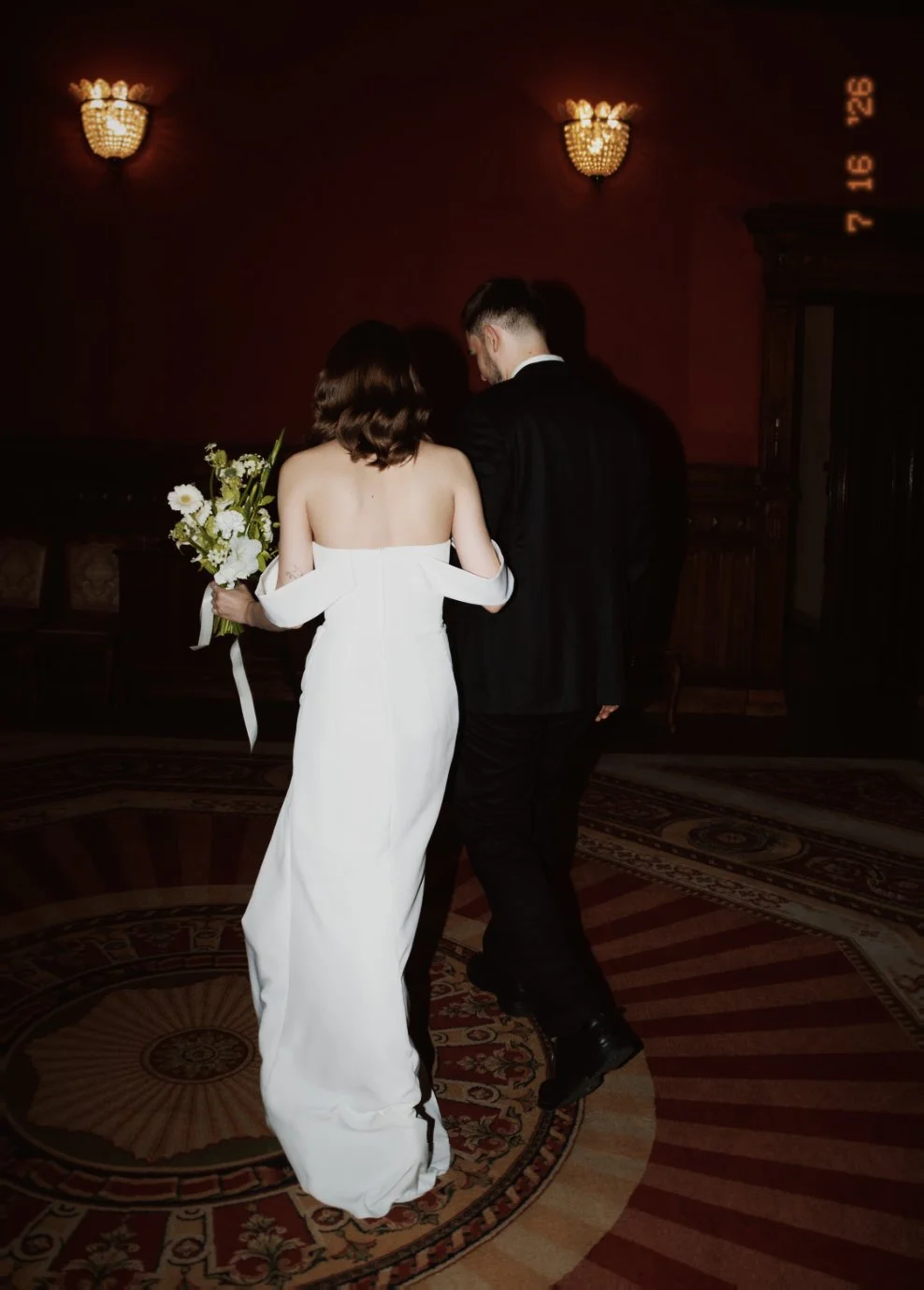 Un couple habillé pour un mariage, la femme en robe blanche et l'homme en costume noir, marche dans une pièce élégante avec un tapis rond au sol, tenant un bouquet de fleurs