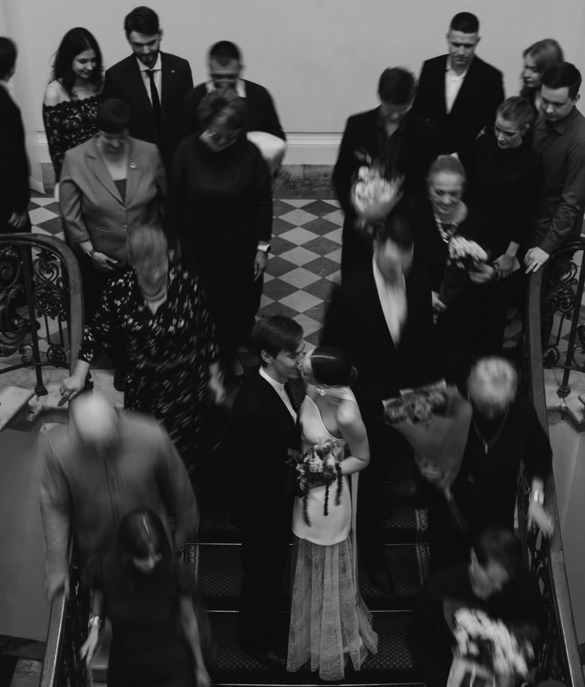 Un groupe de personnes lors d'une célébration, probablement un mariage, avec le couple principal en photo en bas, échangeant un baiser. La scène se déroule sur un escalier en intérieur, en noir et blanc.