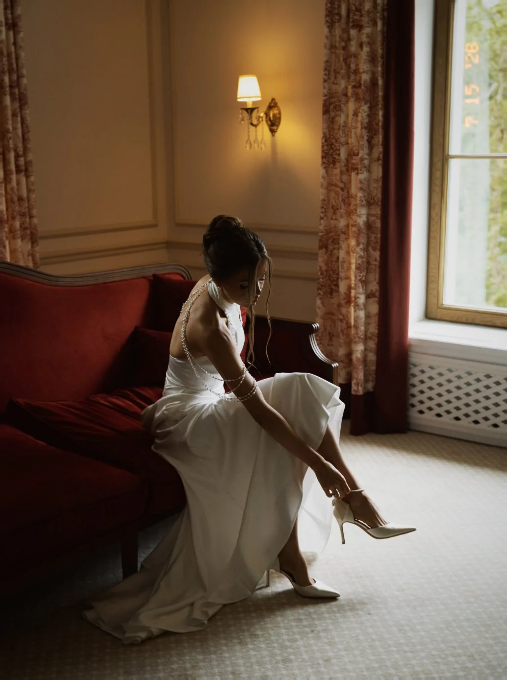 Une femme en robe de mariée assise sur un canapé rouge, en train de mettre ses chaussures à talons blancs dans une pièce aux murs crème, avec une fenêtre laissant entrer de la lumière naturelle.