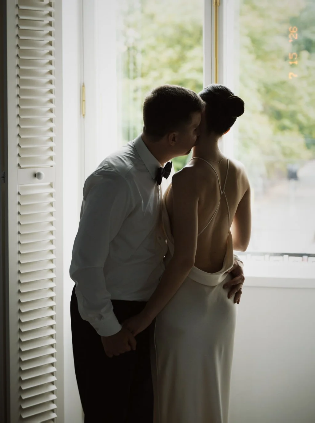 Un couple habillé élégamment, le homme en chemise blanche et nœud papillon, la femme en robe de soirée, partage un moment intime près d'une fenêtre, avec un décor verdoyant visible dehors.