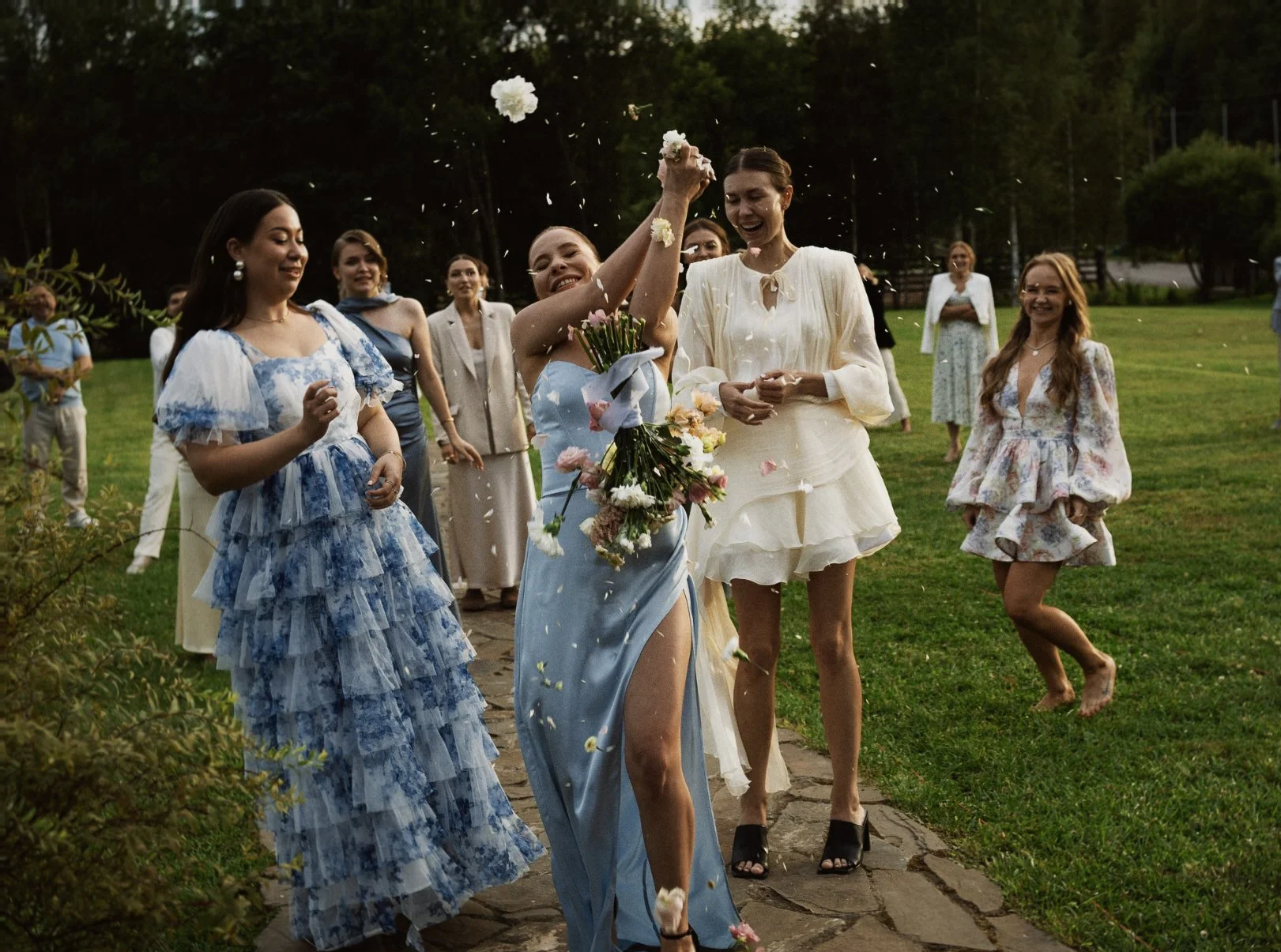 Groupe de femmes, habillées en robes de mariage et de fête, en train de lancer des pétales de fleurs lors d'une célébration en plein air, avec un fond de verdure et de jeunes arbres.