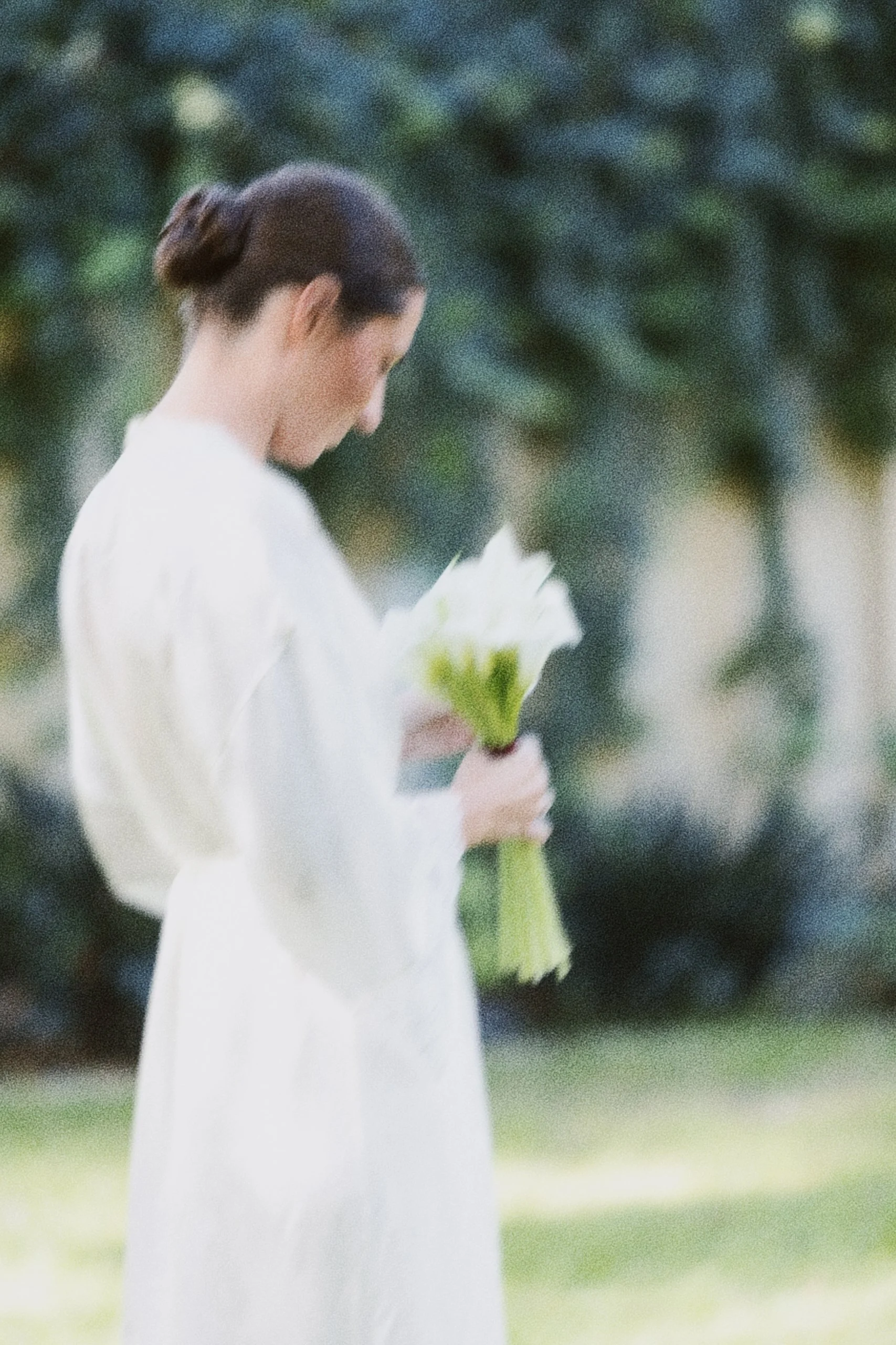 Femme portant une robe blanche, tenant une fleur blanche dans ses mains, en extérieur avec des arbres en arrière-plan.