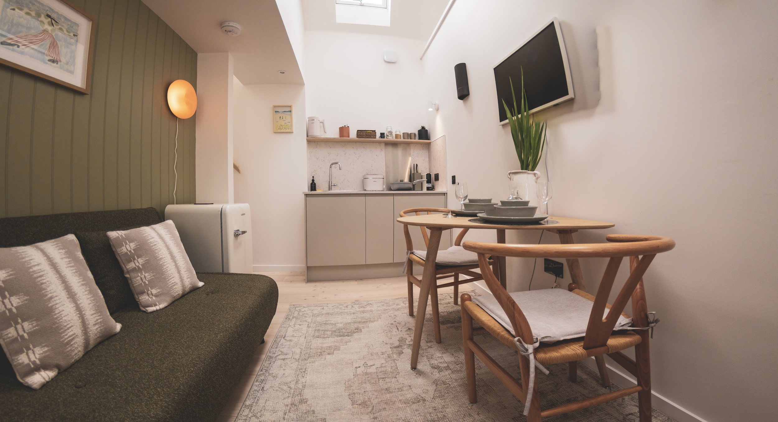 Cozy living space with a sofa with striped pillows, a wooden dining table with matching chairs, a small kitchen area in the background, a wall-mounted TV, and decorative elements like plants and artwork.