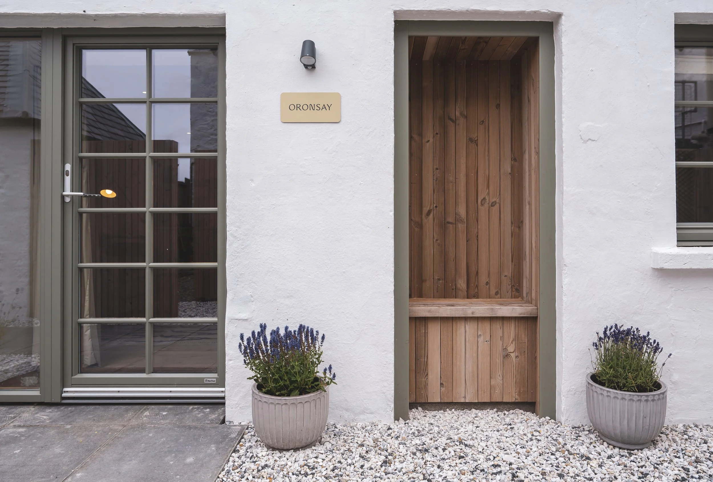 The image shows the exterior of a modern house with a white textured wall, a glass door, and a wooden door with a small step. There are potted lavender plants at the base of the walls and a gravel area in front of the house.