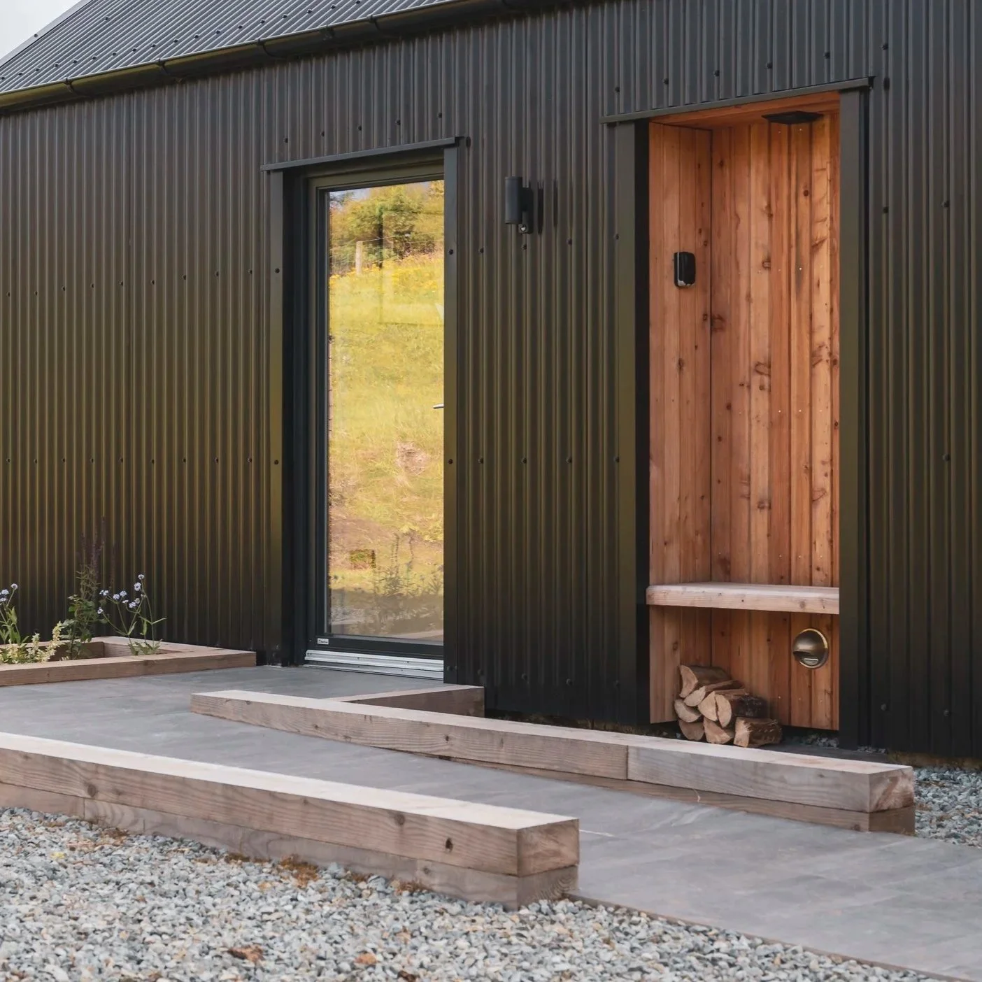 Modern building exterior with black corrugated metal siding, a glass door, and a wooden niche with firewood inside.