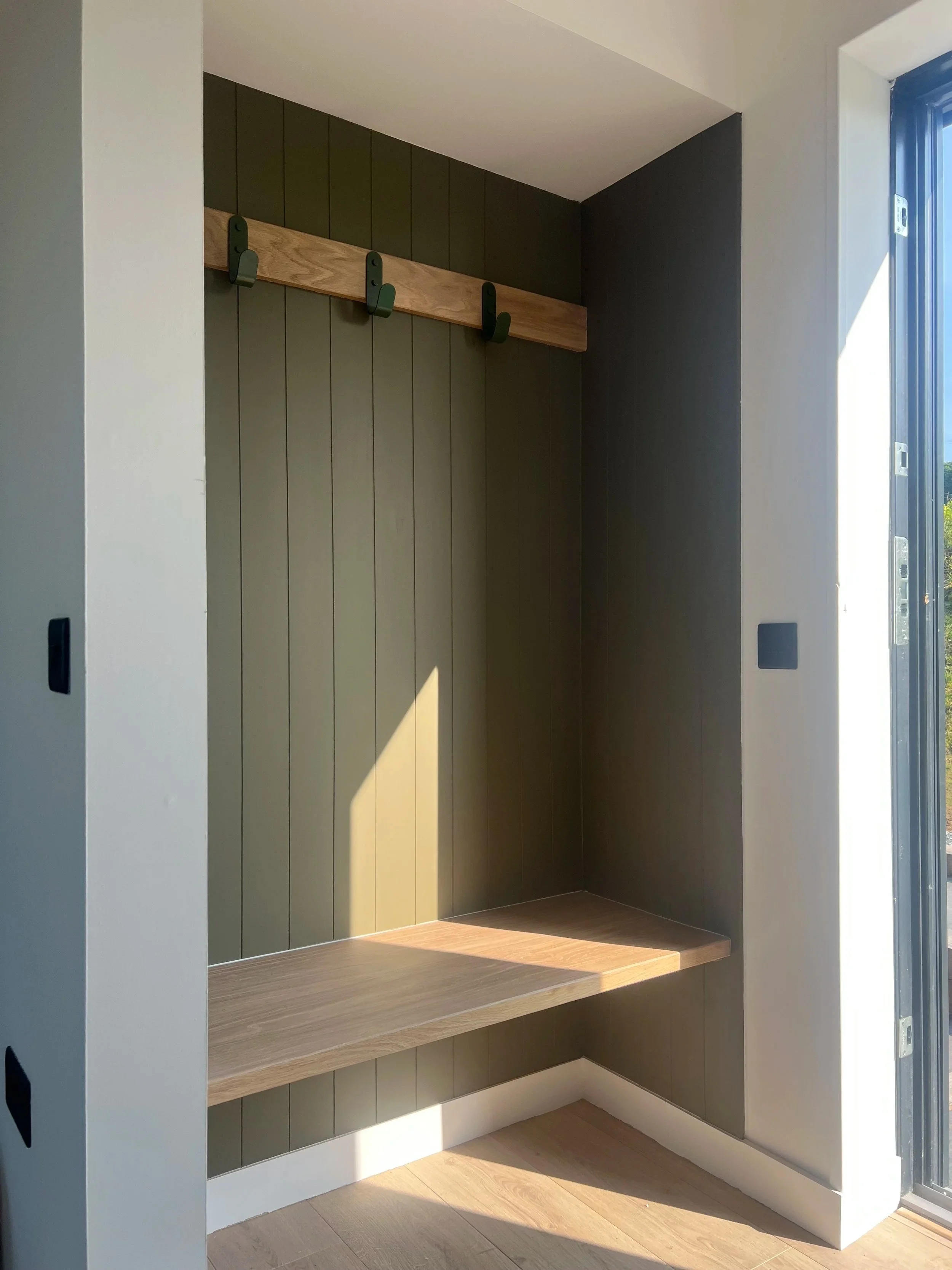 Interior view of a small, modern, built-in closet space with a light wooden bench beneath vertical green panels and a black hook on a wooden rack, bathed in natural sunlight from a nearby glass door.