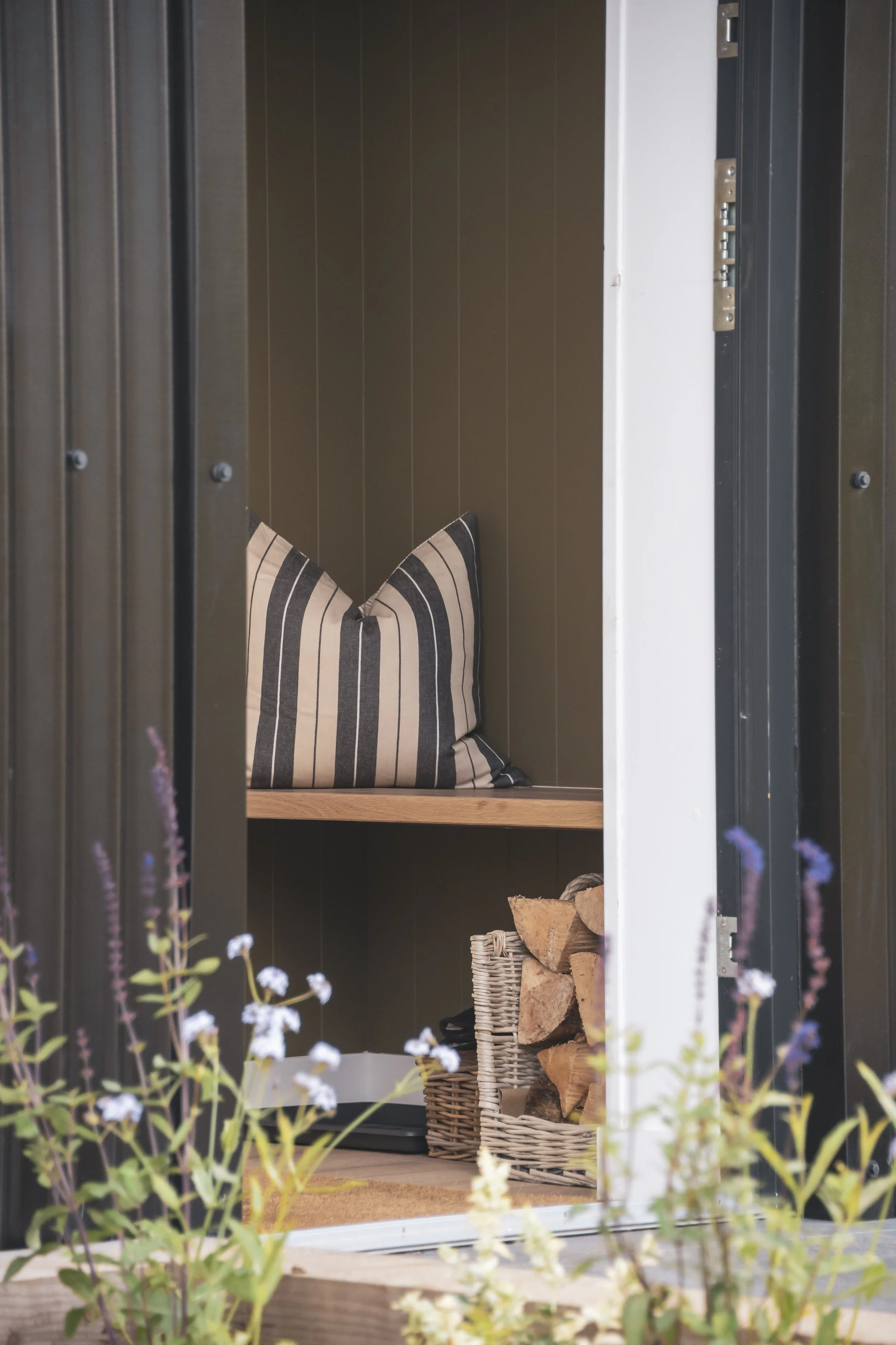 Inside a small storage shed with wood panel walls, a striped pillow, a basket of chopped firewood, and a black dishwasher.