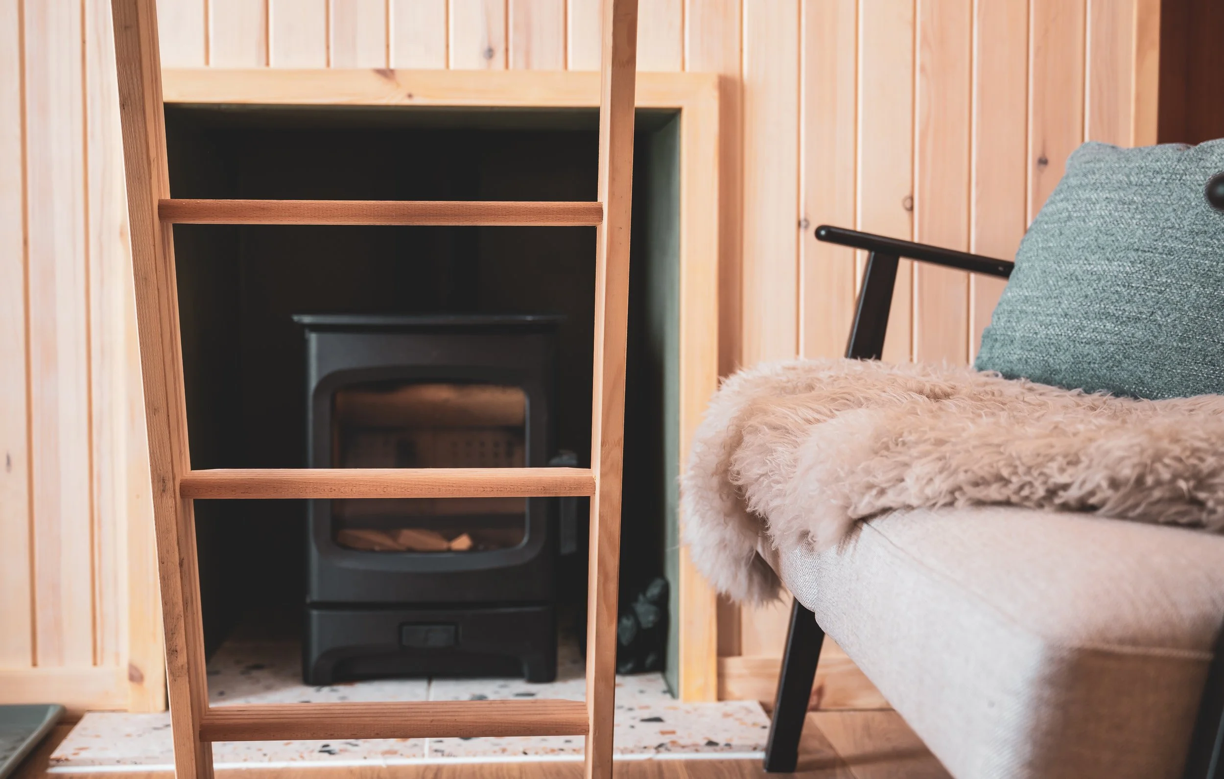 A cozy interior corner with a wooden wall, a plush beige blanket on a white couch with a textured green pillow, and a small black wood stove in a wooden housing unit.