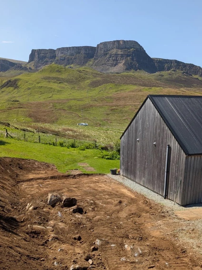 A hillside with a grassy slope and rocky cliffs in the background, a black shed on the right, and a patch of dirt in the foreground, with a car parked on the distant road.