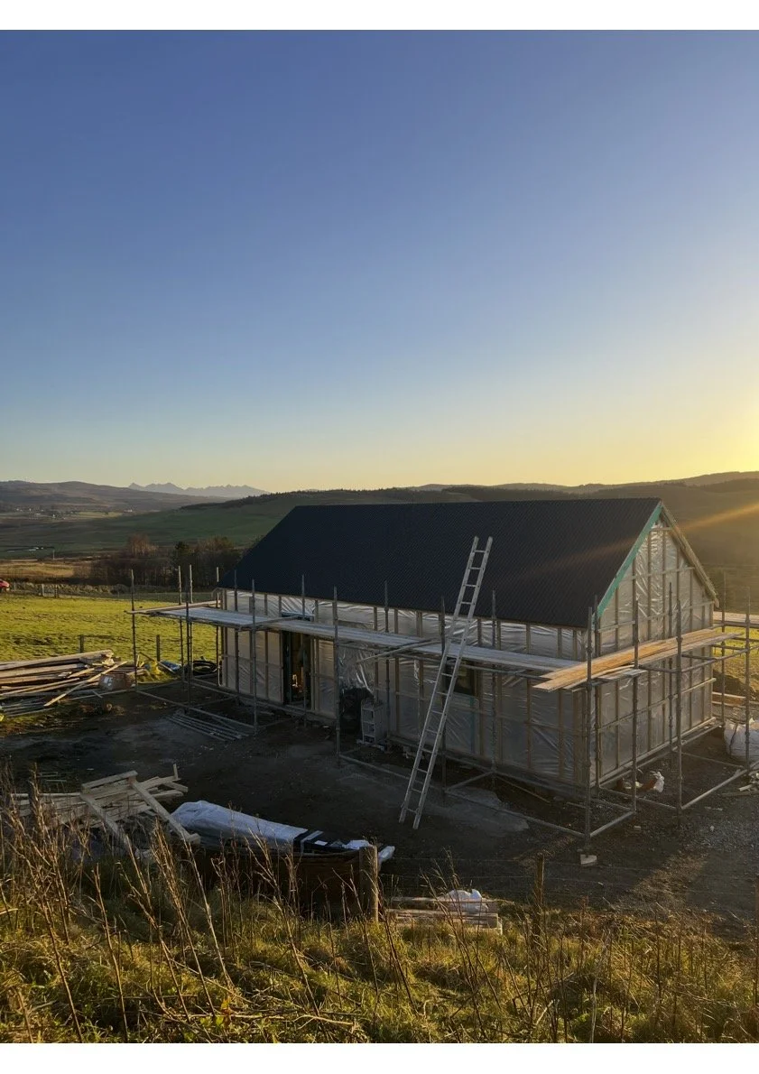 A house under construction in a rural area at sunset, with scaffolding surrounding it and a ladder leaning against the roof.