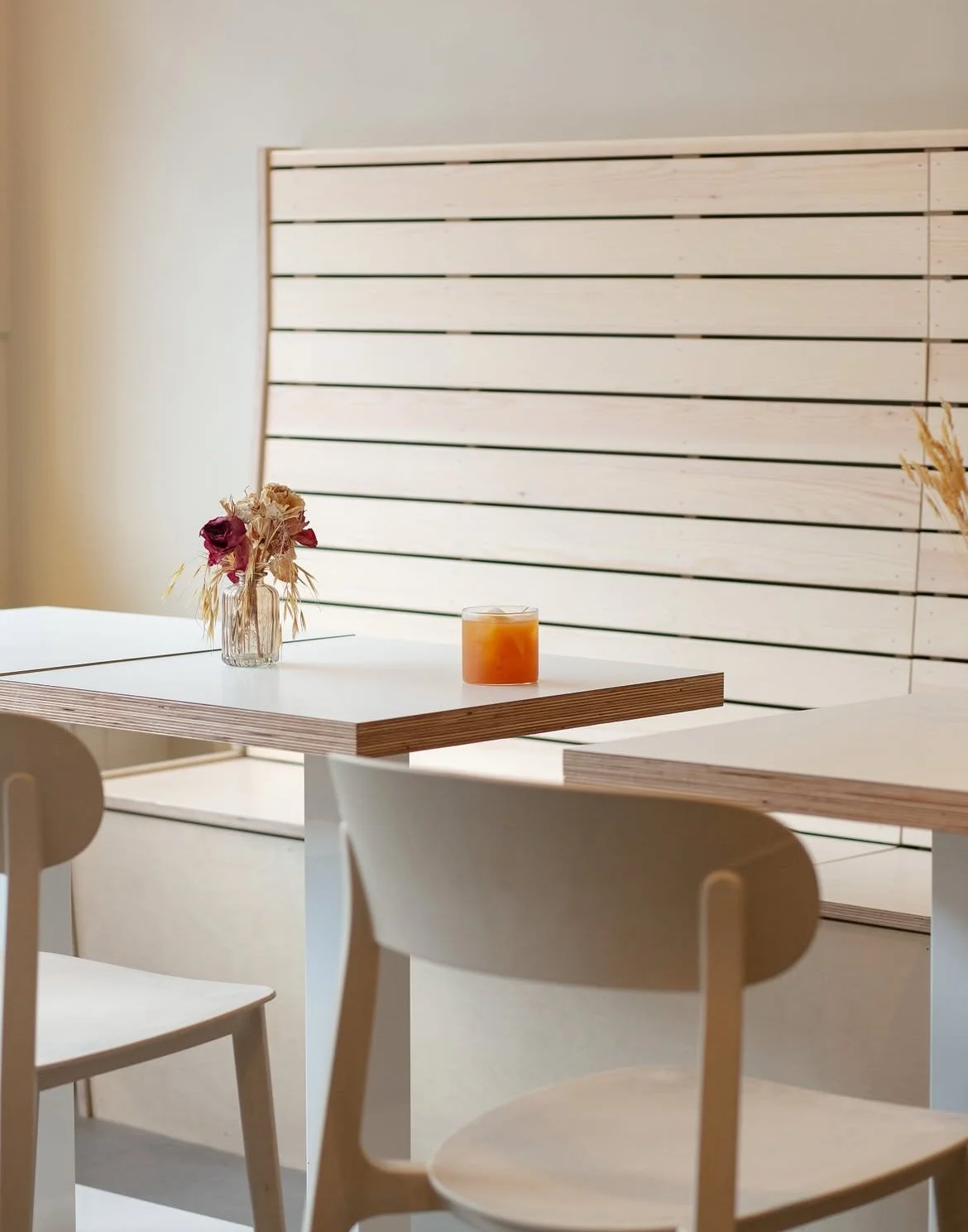 A minimalistic dining area with a white table, beige chairs, a dried flower arrangement in a glass vase, and a candle on the table. In the background, there is a large light wooden slatted wall.