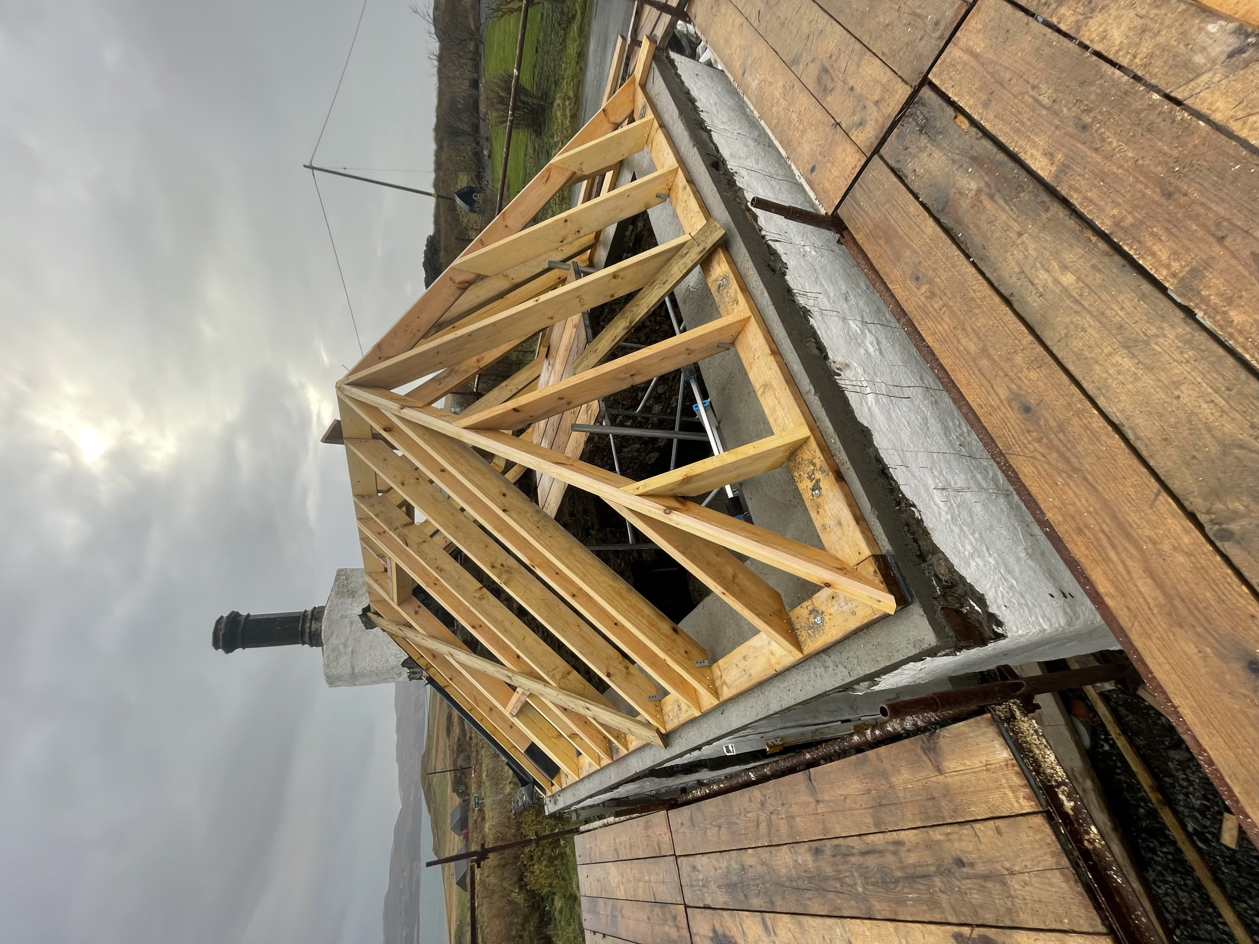Construction site with wooden framing on top of a concrete base, adjacent to a partially visible rooftop with shingles and a chimney, overcast sky in the background.