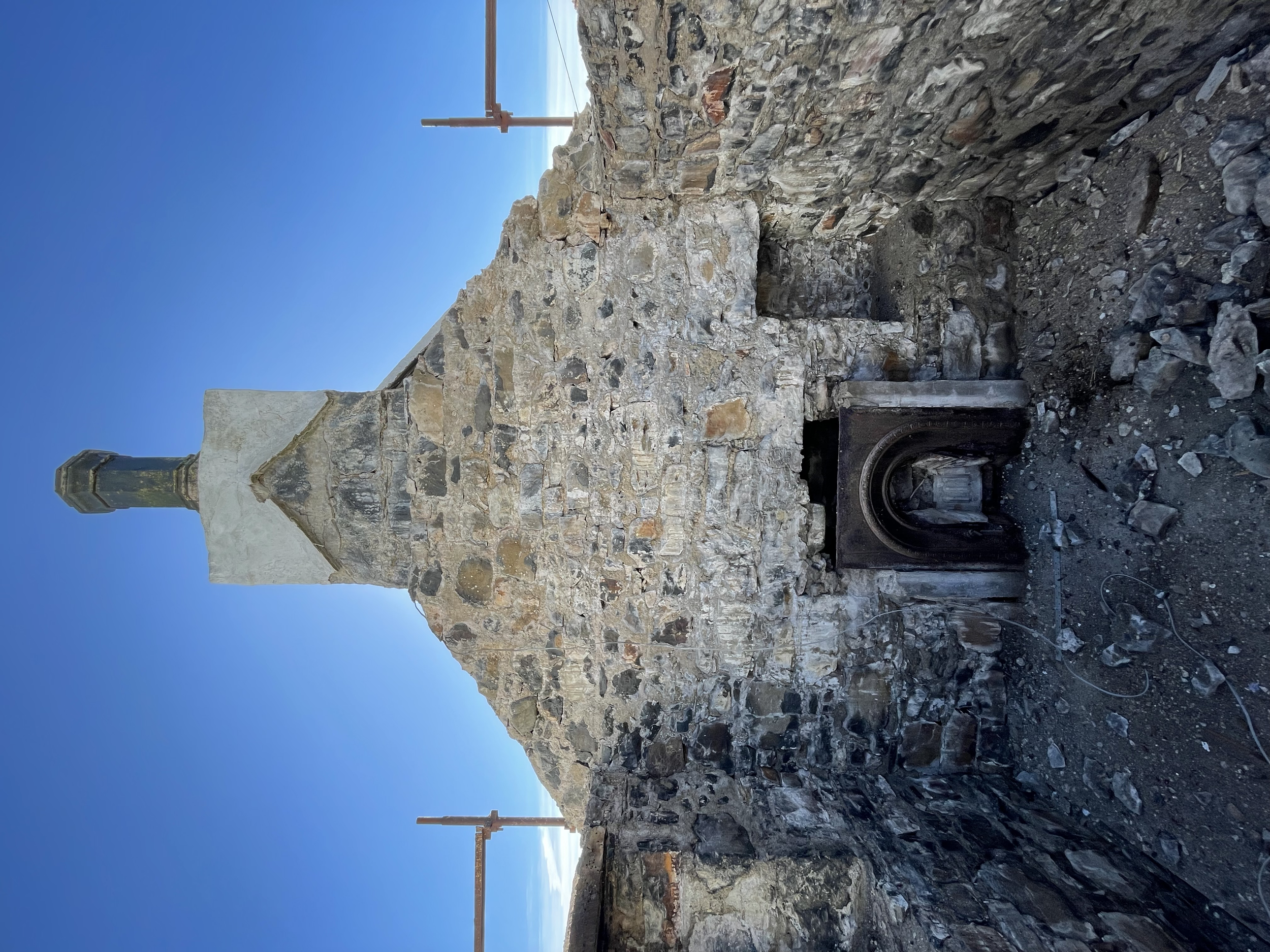 View of an old stone structure with a metal door, some rusted pipes, and a clear blue sky in the background.