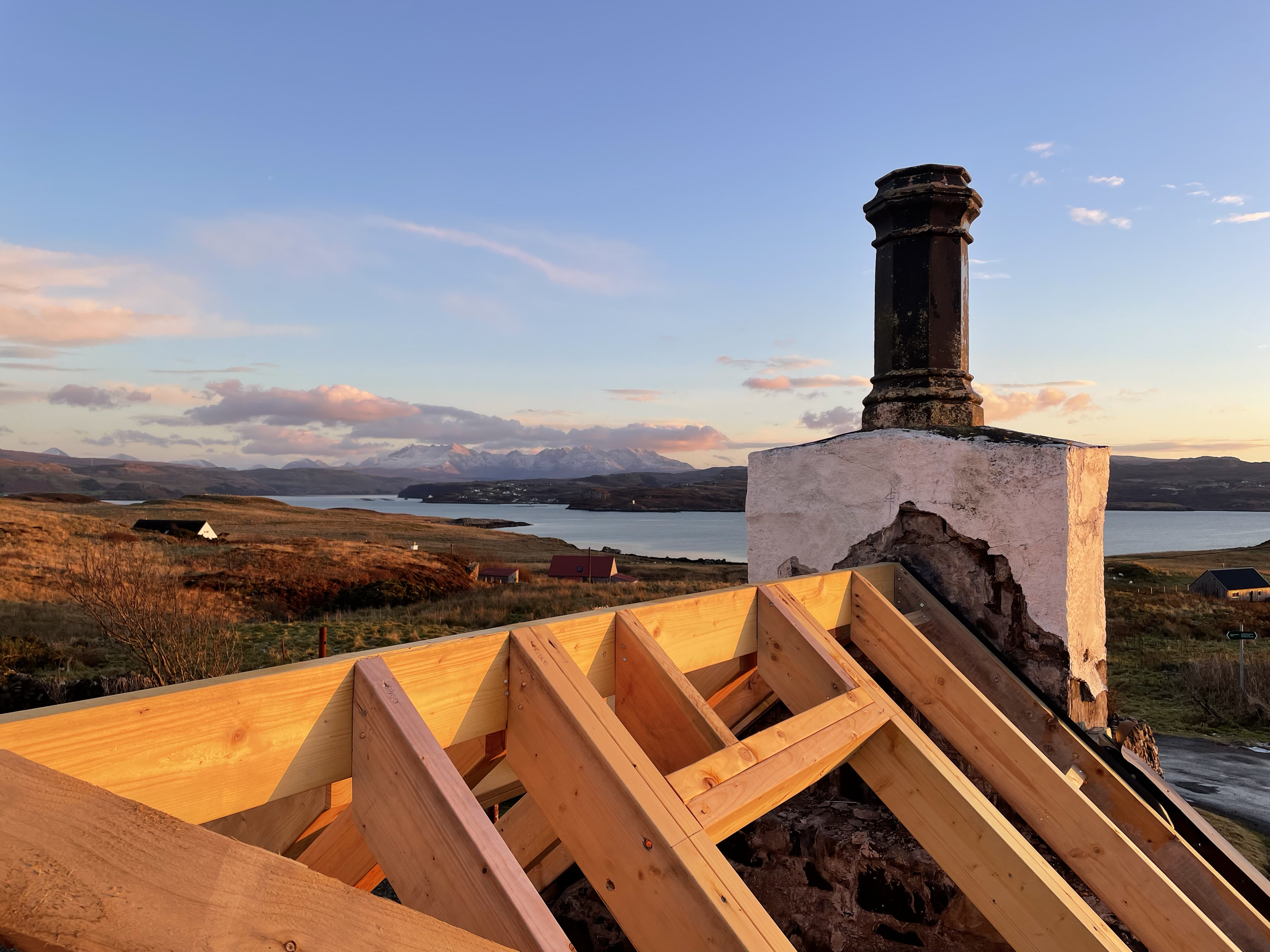 A house roof under construction with wooden framework, an old chimney, and a scenic background of lakes, hills, and mountains at sunset.