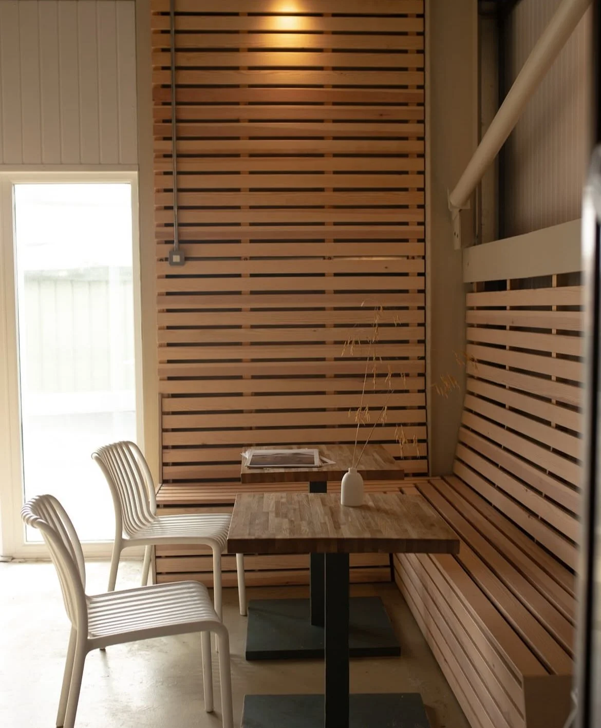 Interior of a modern room with slatted wood walls, a wooden table with a vase and dried grass, and white chairs.
