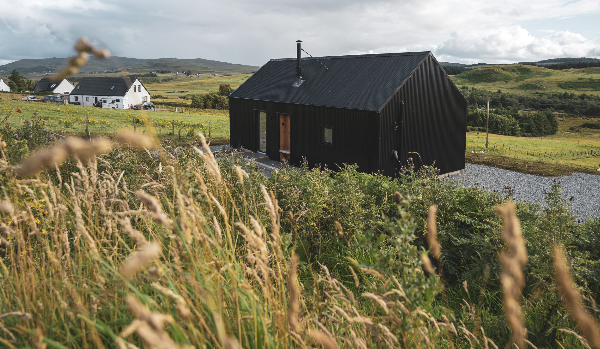A black modern house with a sloped roof and minimal windows in a rural landscape, surrounded by tall grass and wildflowers, with other houses and rolling hills in the background under cloudy skies.