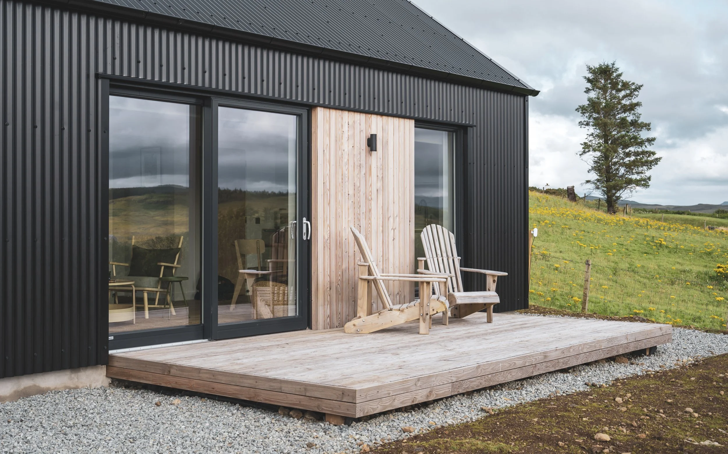 A modern house with a black metal exterior and wooden accents, featuring a small outdoor deck with wooden chairs, set in a rural landscape with rolling hills, wildflowers, and a cloudy sky.