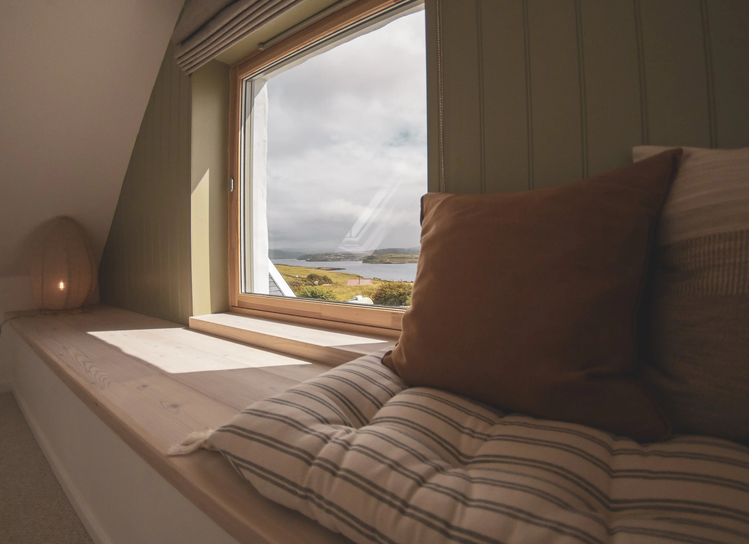 A cozy bedroom with a large window showcasing a scenic view of a cloudy sky, water, and green hills. The bed is made with striped sheets and a brown pillow, and there is a small bedside lamp.
