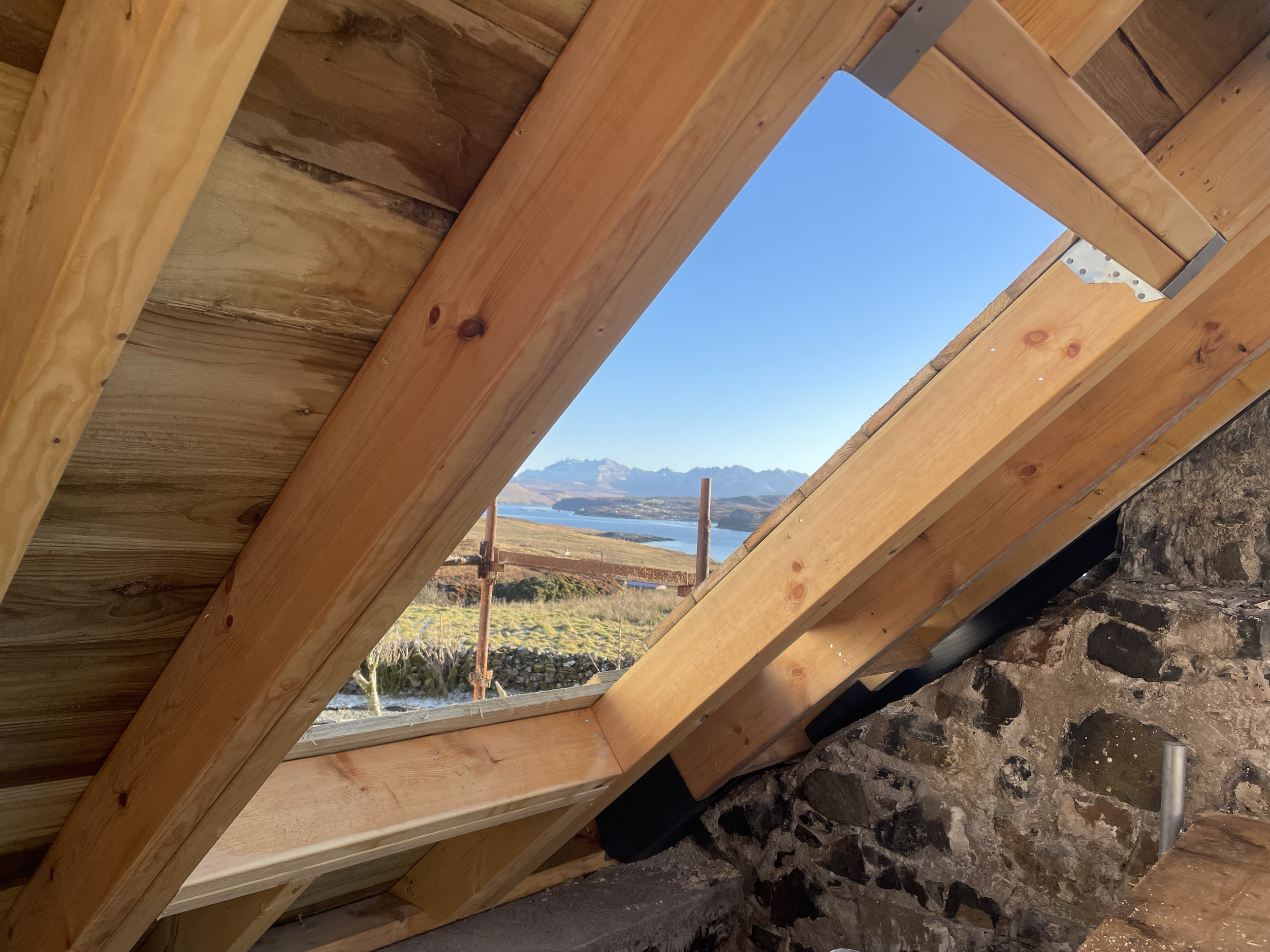 View through a rectangular opening in the wooden roof structure, revealing mountains, a body of water, and a clear blue sky in the distance.