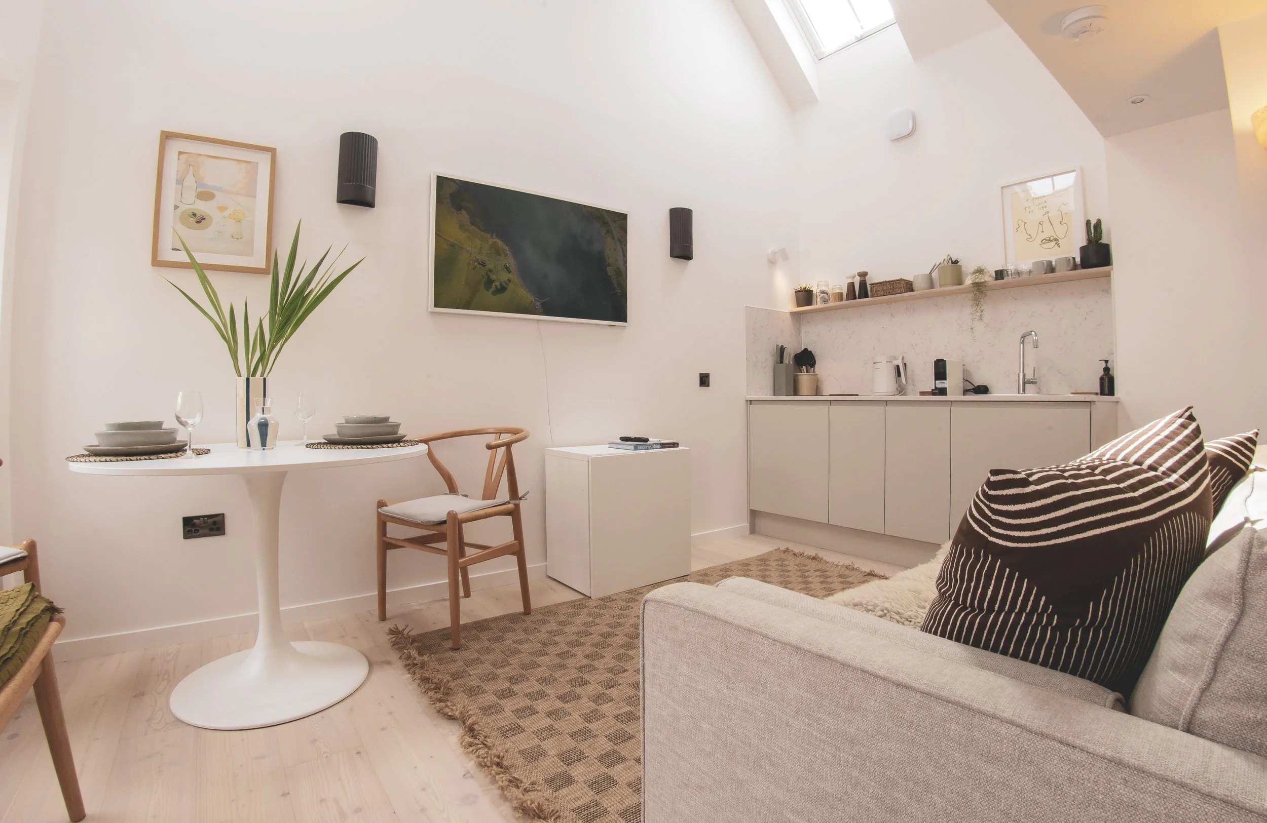 A modern, minimalistic living space with white walls and ceiling, a small dining table with place settings, a wooden chair, a beige sofa with striped pillows, a small kitchenette with cabinets, a sink, and kitchen appliances, wall art, and potted pla
