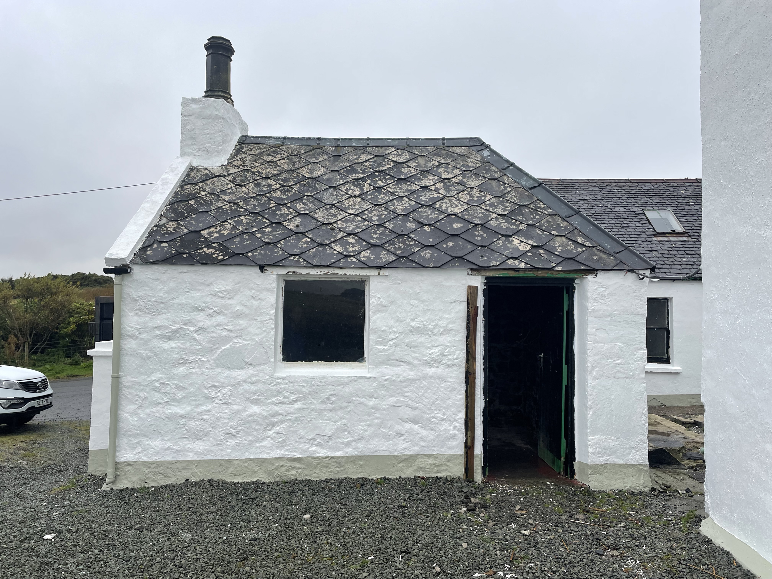 A house with white walls and a dark, slate tile roof showing signs of moss and weathering. There is an open doorway and a window, with a partially built or repaired section. The sky is overcast, and there is a gravel area in front of the house, with 