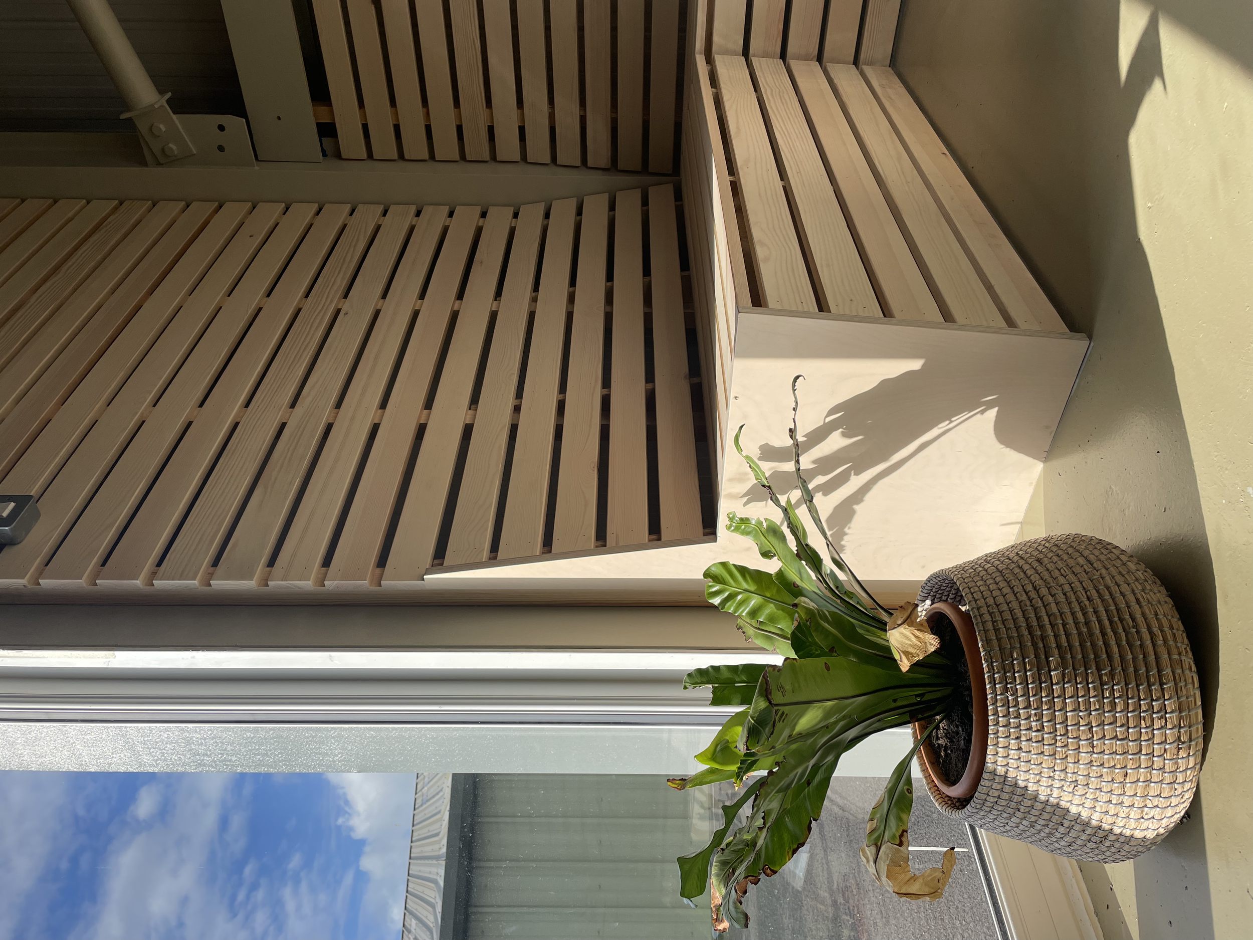 Indoor balcony with a wooden lattice railing, a potted plant with large green leaves, and a wicker basket near the plant. The balcony overlooks a view of the sky and neighboring buildings.