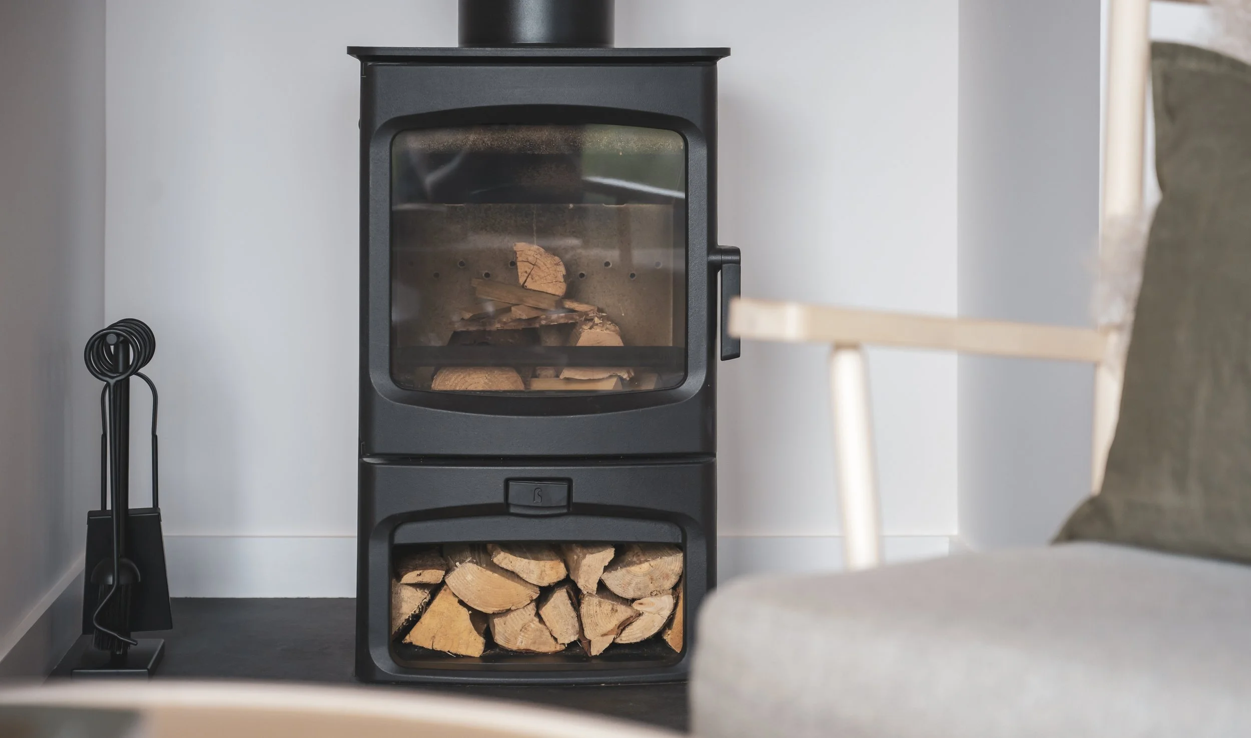 A black wood stove with glass door, filled with firewood inside and stored below, placed against a white wall with a black tool holder and poker off to the side.