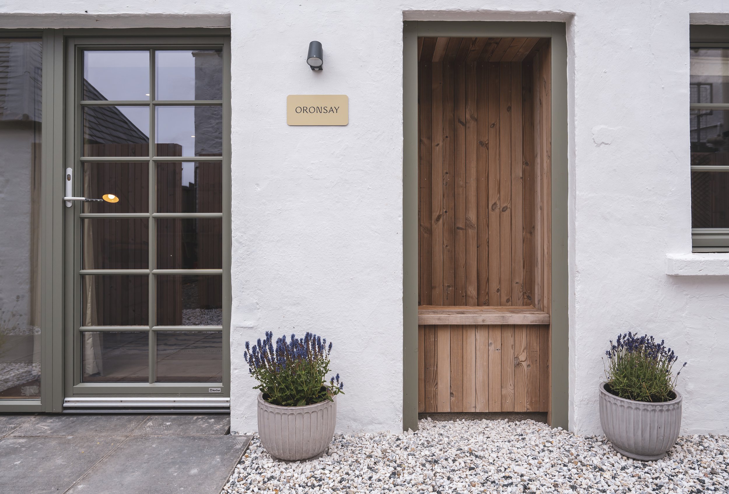 Front of a white house with a glass door on the left and a wooden door on the right, two potted lavender plants in front, and a small gravel garden area.