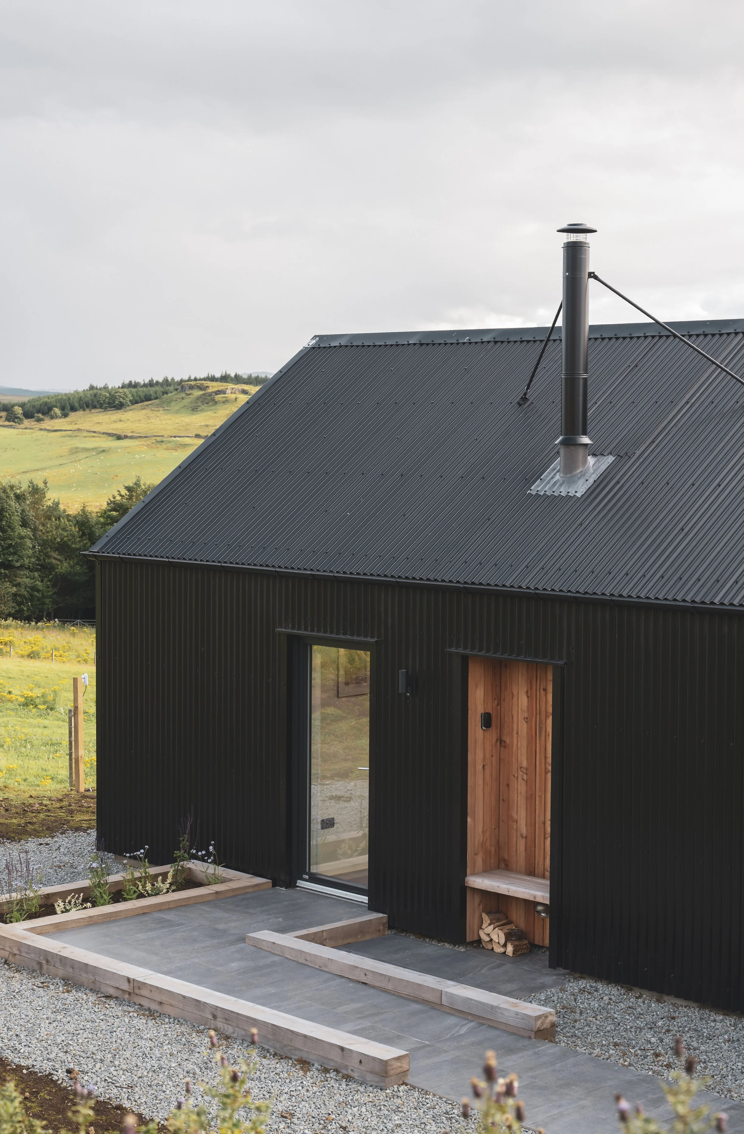 A modern black metal house with a sloped roof and a chimney. The front has a glass door and a small wooden porch. There is a garden bed and a pathway leading to the house, with a rural landscape of rolling hills and fields in the background.