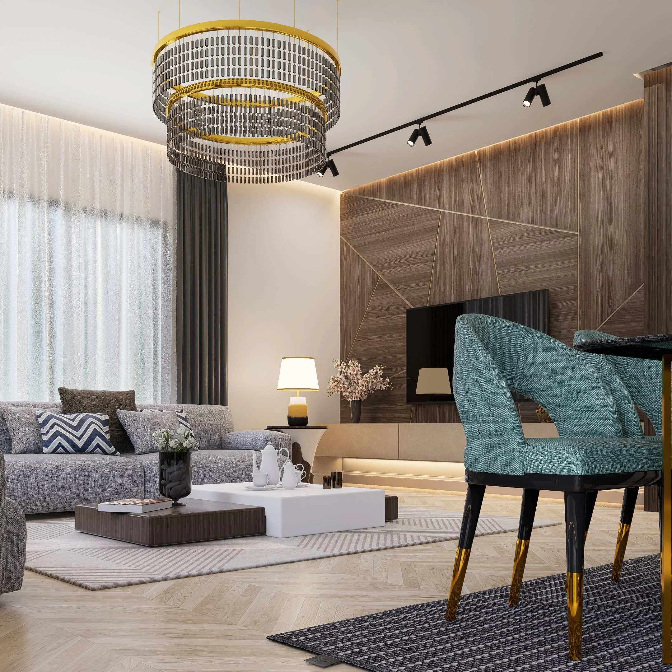 Modern living room with a large decorative chandelier hanging from the ceiling, a grey sectional sofa with patterned cushions, a white coffee table with decorative objects, a beige rug, and wooden wall paneling with integrated lighting. There are also teal dining chairs with black legs, black track lighting, and a window with white sheer curtains and dark drapes.