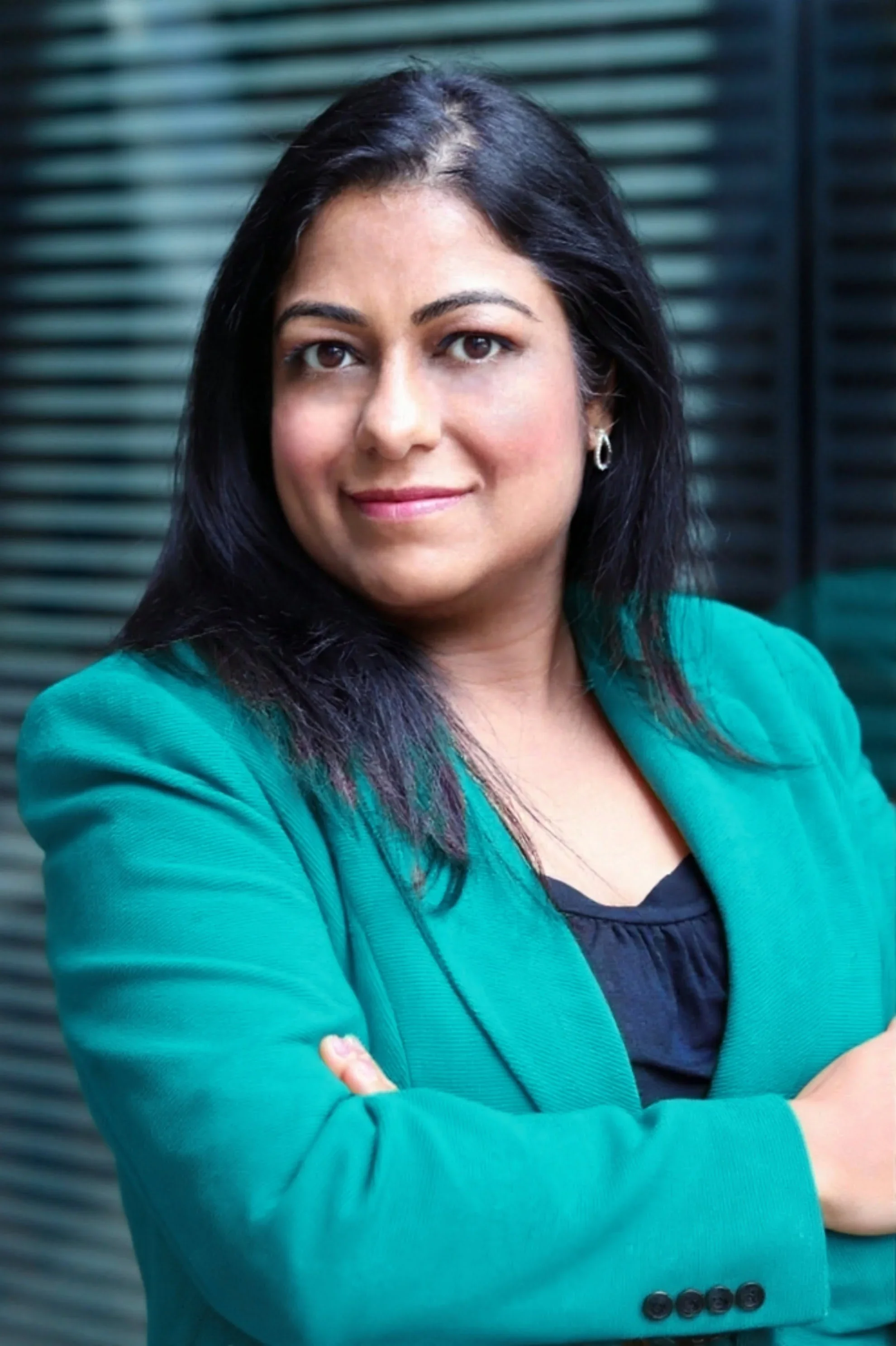A woman with dark hair wearing a teal blazer and black top, standing with arms crossed in front of a dark, slatted background.