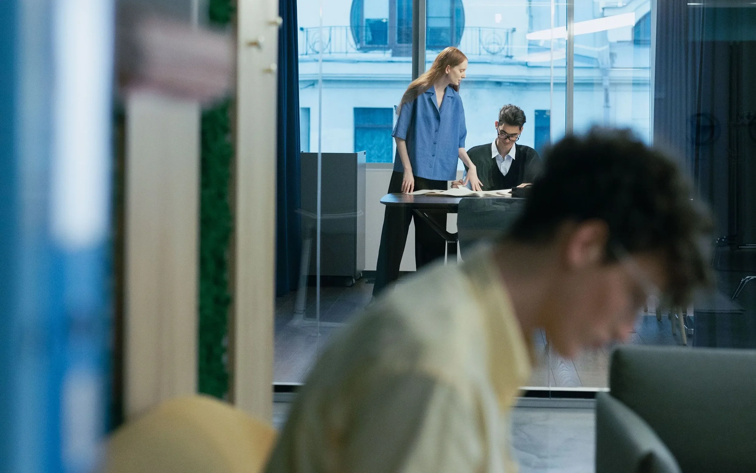 Two women in a modern office, one standing and the other seated at a table, with large windows showing buildings outside.