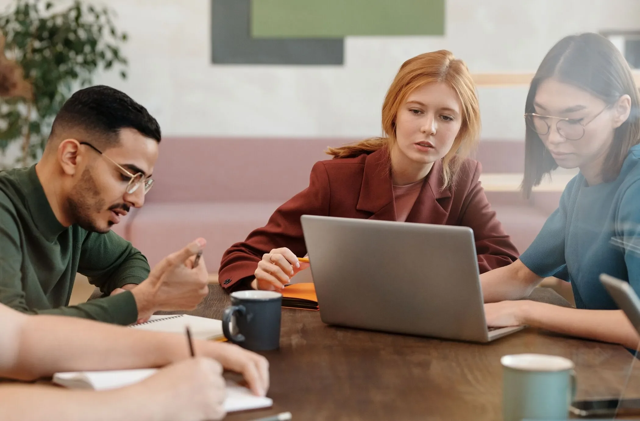 Three people sitting at a table during a meeting, looking at a laptop. One is using a smartphone, another has a notebook, and a third is pointing at the laptop. Coffee mugs and notebooks are on the table.