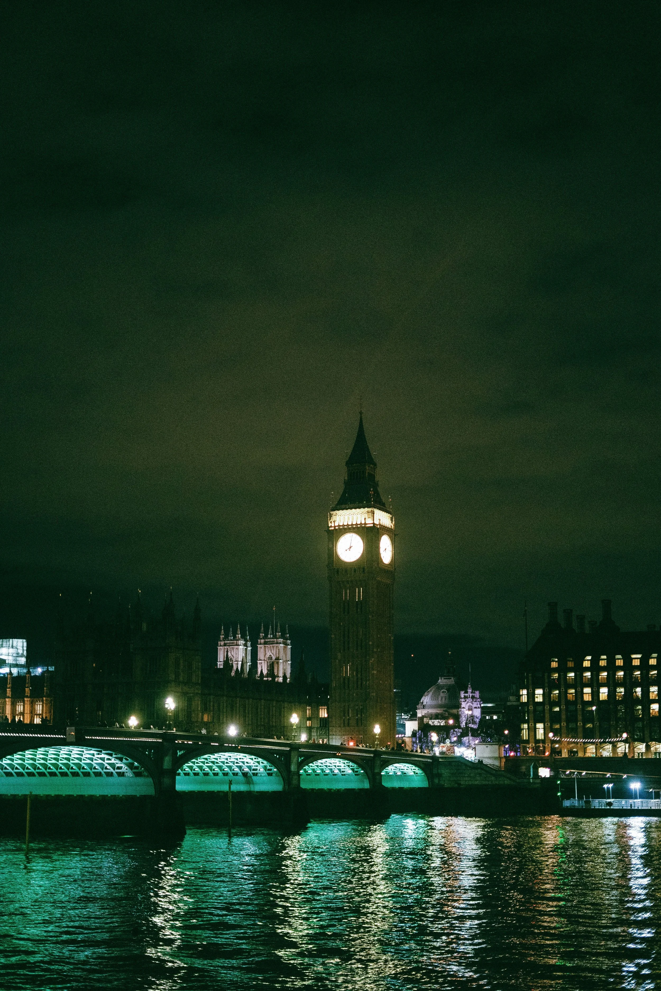 Nighttime view of Big Ben clock tower in London, illuminated, with reflections on the River Thames and city buildings in the background.