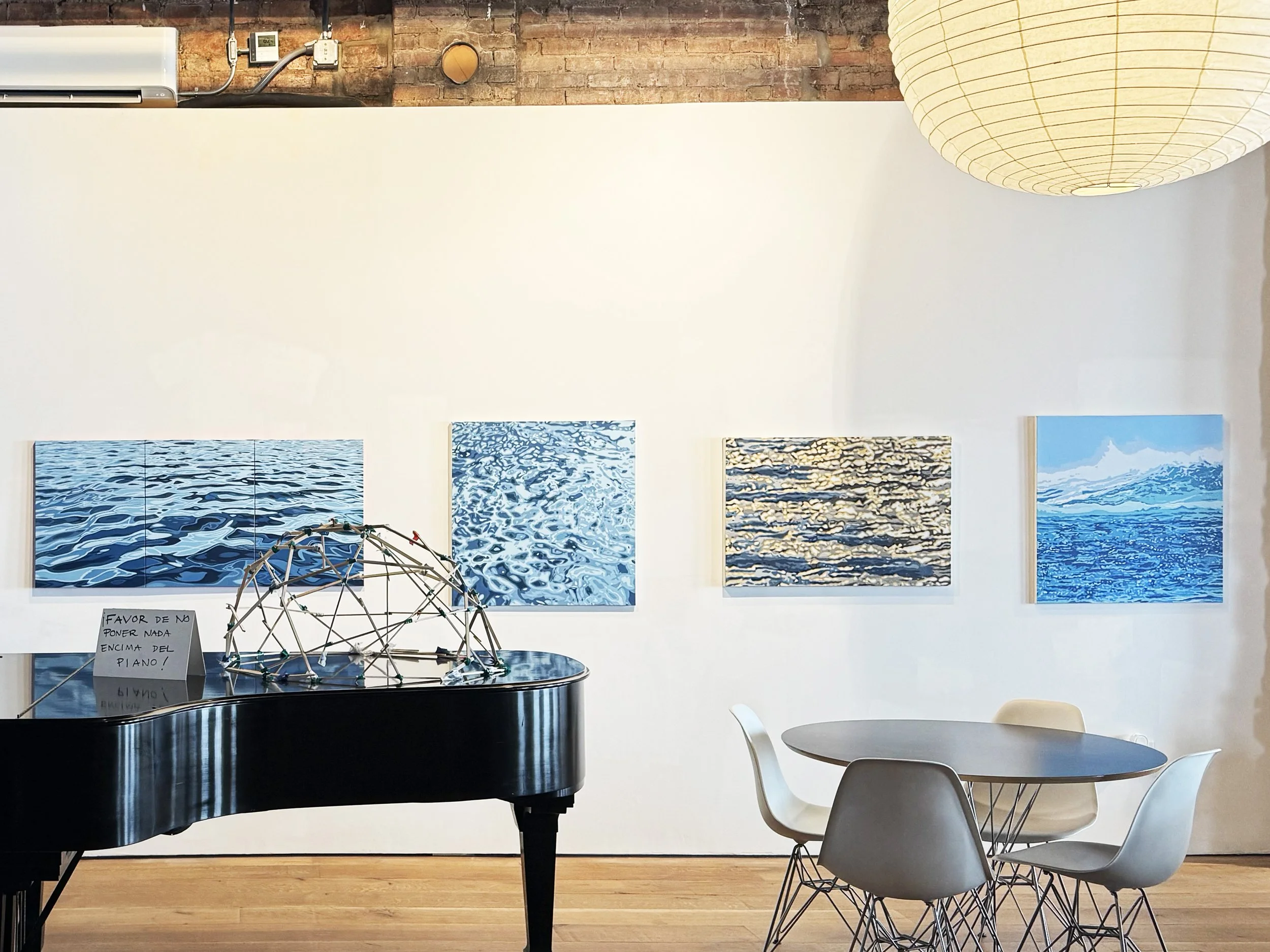 An indoor art gallery with abstract blue watercolor paintings on the white wall, a black grand piano in the foreground with a note that says 'Favor de no poner nada encima del piano!', a round white table with four white chairs, a large round paper lantern light, an exposed brick wall, and plants in the background.
