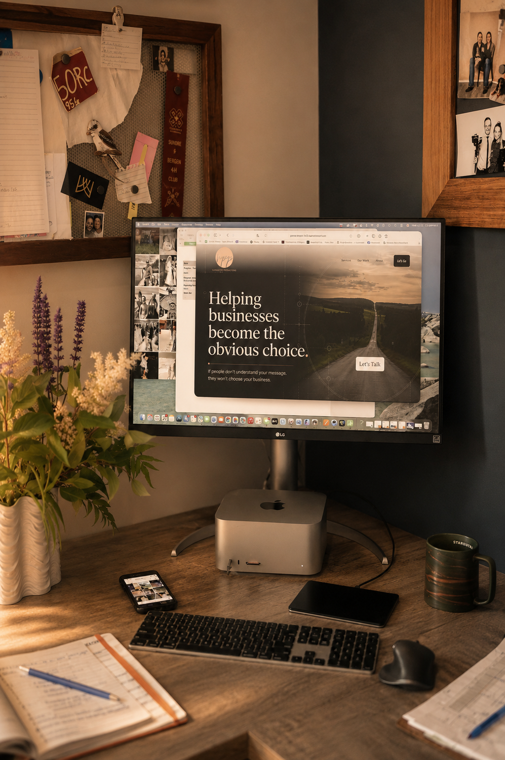 A desk setup with a large monitor displaying a website that says "Helping businesses become the obvious choice." The desk has a keyboard, mouse, smartphone, notebook, pen, a black book, a coffee mug, and a plant. There are two cork boards on the wall behind the desk with various photos, notes, and decorations.