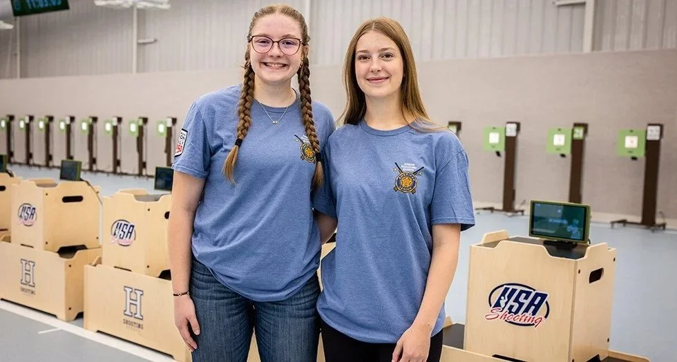 Two young women in blue shirts with shooting team logos, smiling, standing in an indoor shooting range with computer targets and shooting stations in the background.