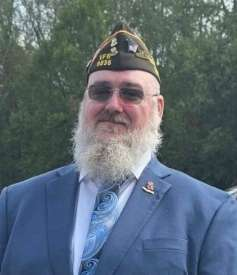 A man with a white beard and glasses, wearing a military-style cap and a blue suit, standing outdoors with trees in the background.