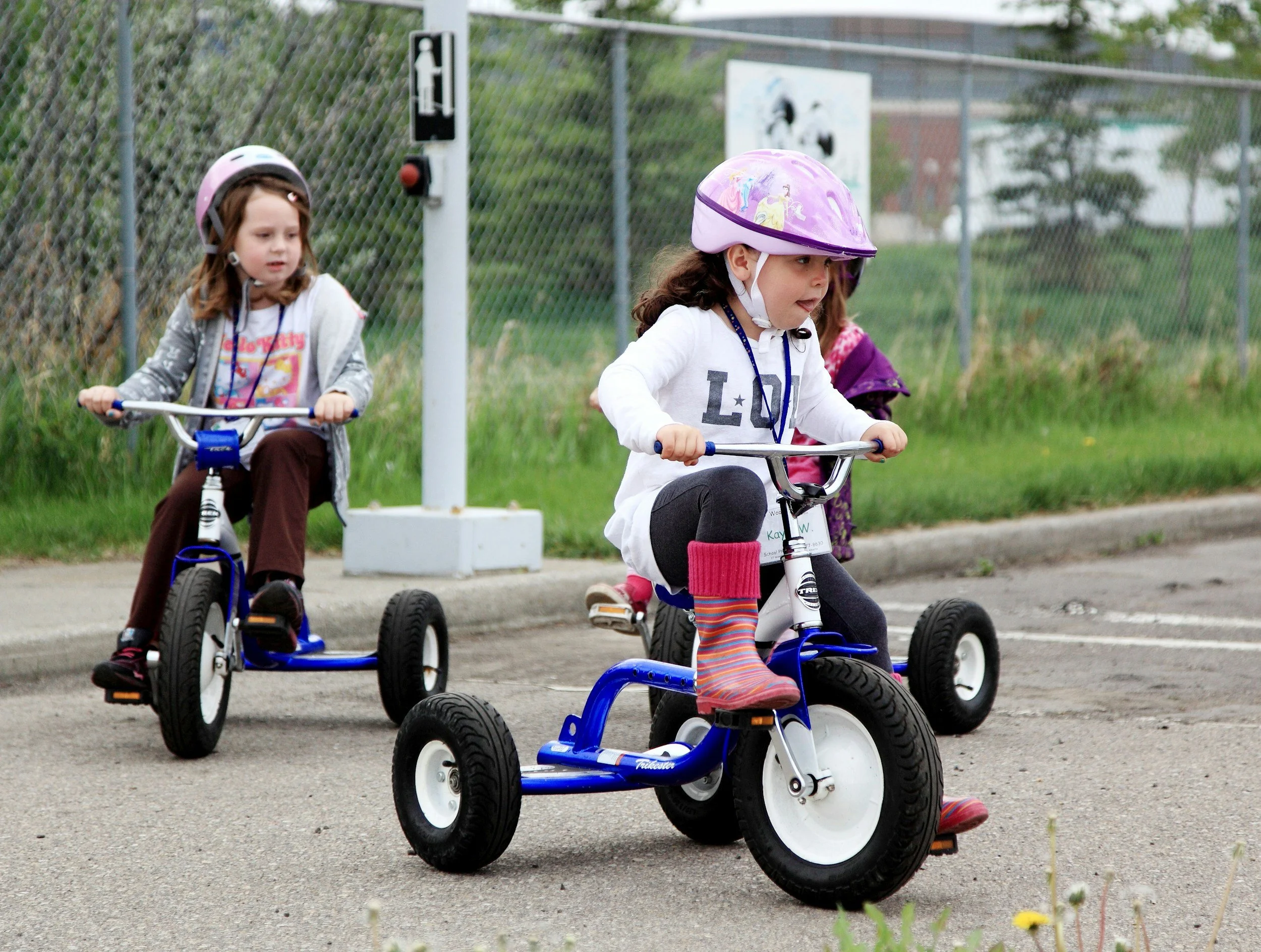 Three young girls riding tricycles outdoors near a chain-link fence during daytime. The girl in the foreground is wearing a pink helmet, a white shirt with writing, and colorful striped socks. Another girl behind her has a purple helmet and is wearing a gray jacket. The third girl is partially visible in the background.
