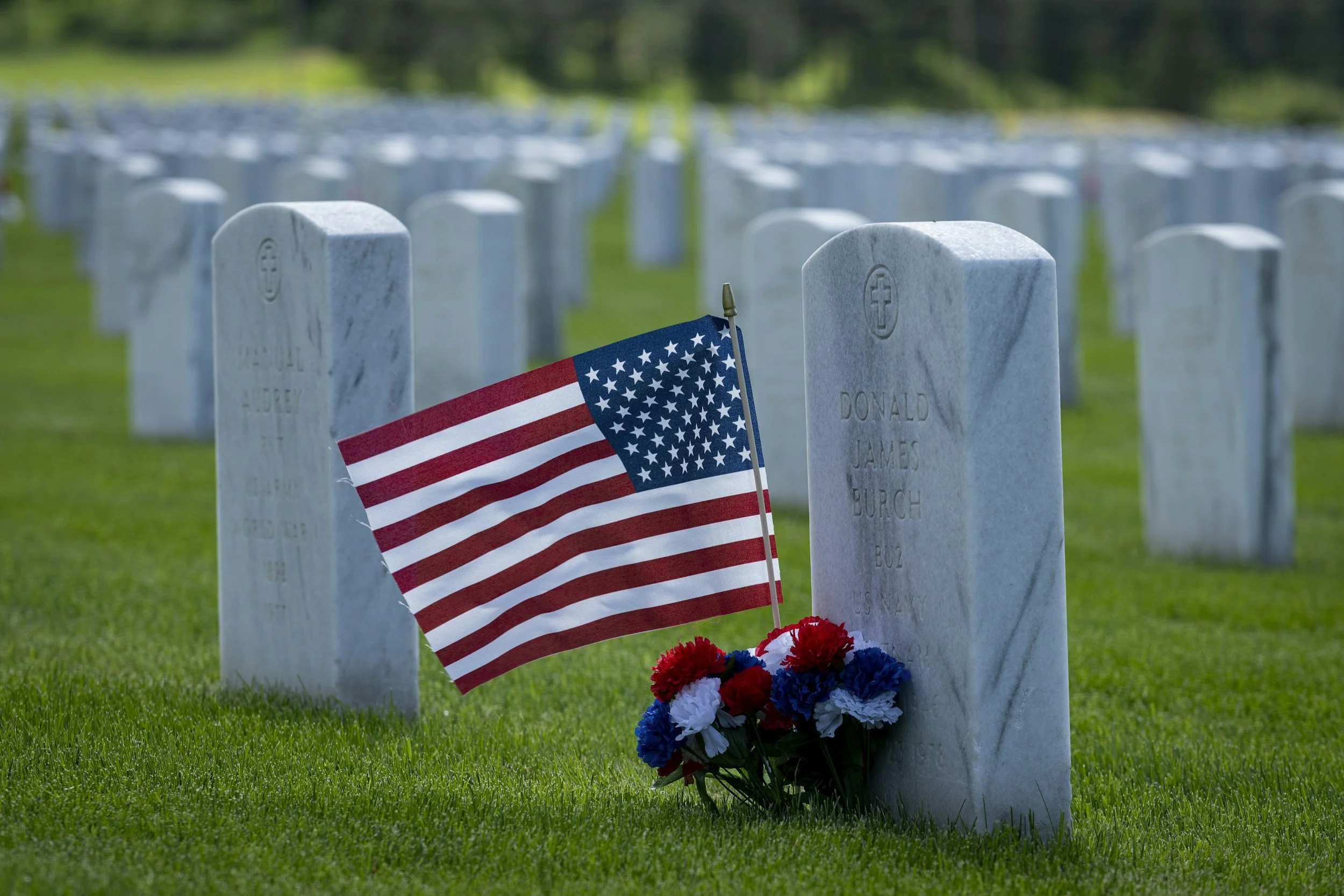 A row of white grave markers in a cemetery, with an American flag and flowers placed in front of one, under a green lawn and trees.