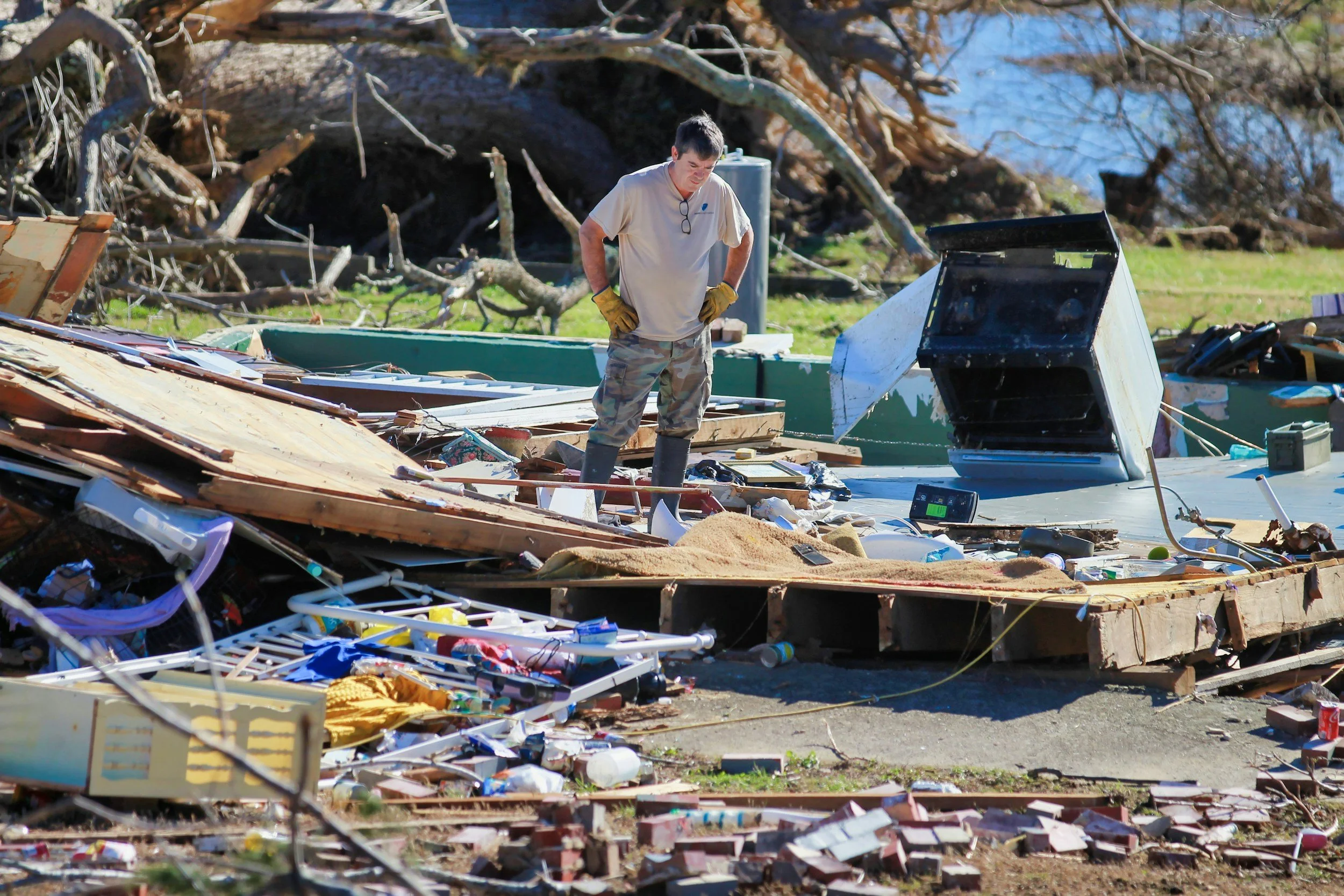 A man standing among debris and wreckage after a storm or disaster, looking down at the destruction, with trees and a body of water in the background.