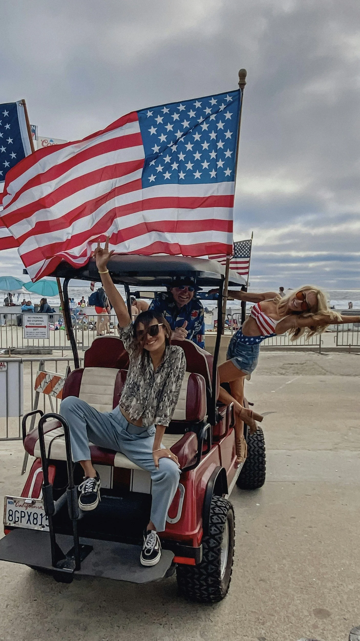 Three people on a golf cart decorated with American flags at the beach. One woman sits on the cart, another woman leans out of the cart, and a man is seated behind them all, smiling and wearing sunglasses.
