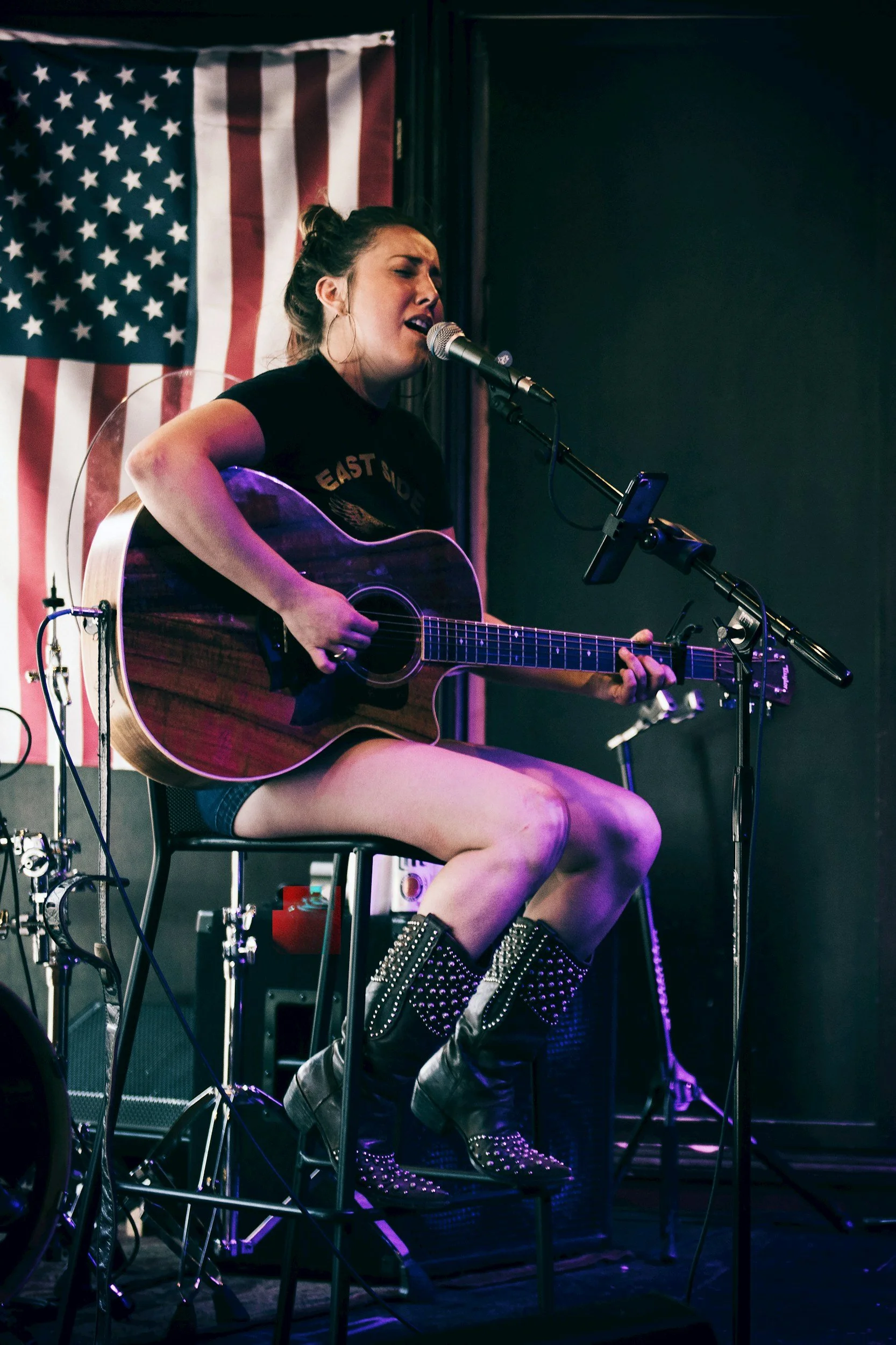 A woman with dark hair tied up, singing into a microphone while playing an acoustic guitar on stage. She wears hoop earrings, a black t-shirt with text, shorts, and studded boots. An American flag hangs behind her, and the stage has various equipment and purple lighting.